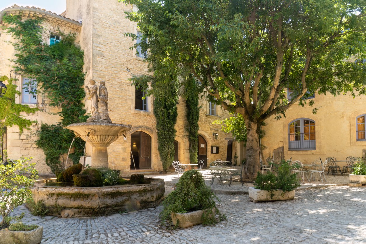 Courtyard with a fountain, a large tree, outdoor tables and chairs, and a beige stone building with windows and greenery.