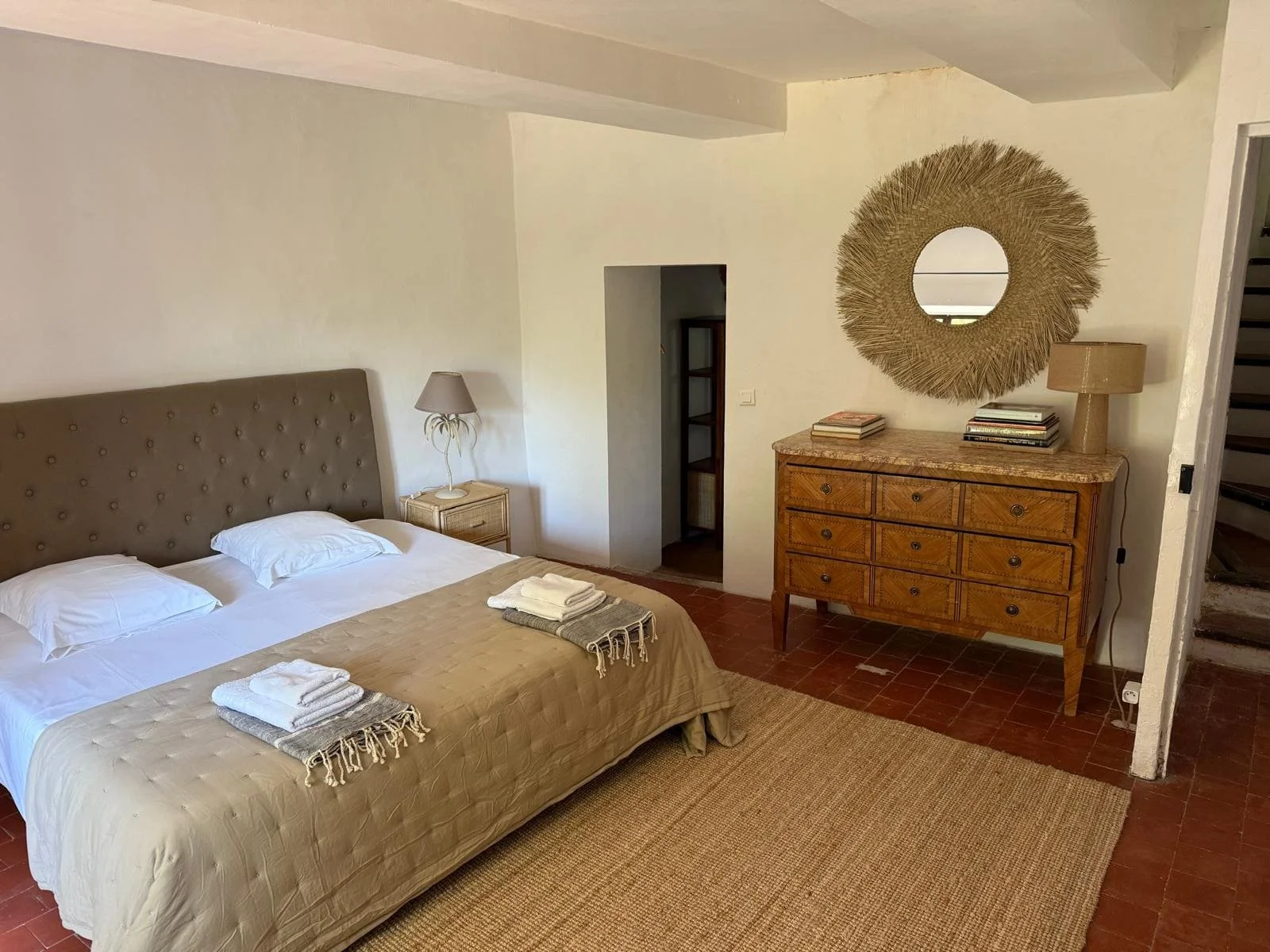 Chambre avec un lit, deux oreillers, une lampe de chevet, une commode en bois avec des livres et un miroir rond en décoration.