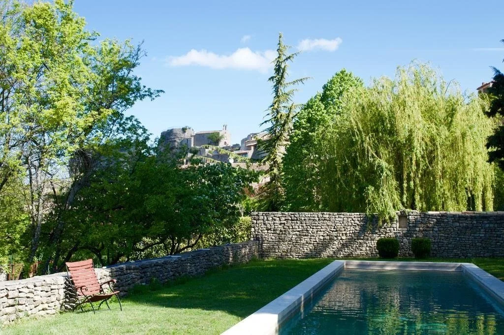 Jardin avec piscine, mur en pierre, banc en bois, arbres verts et château au loin sous un ciel bleu.
