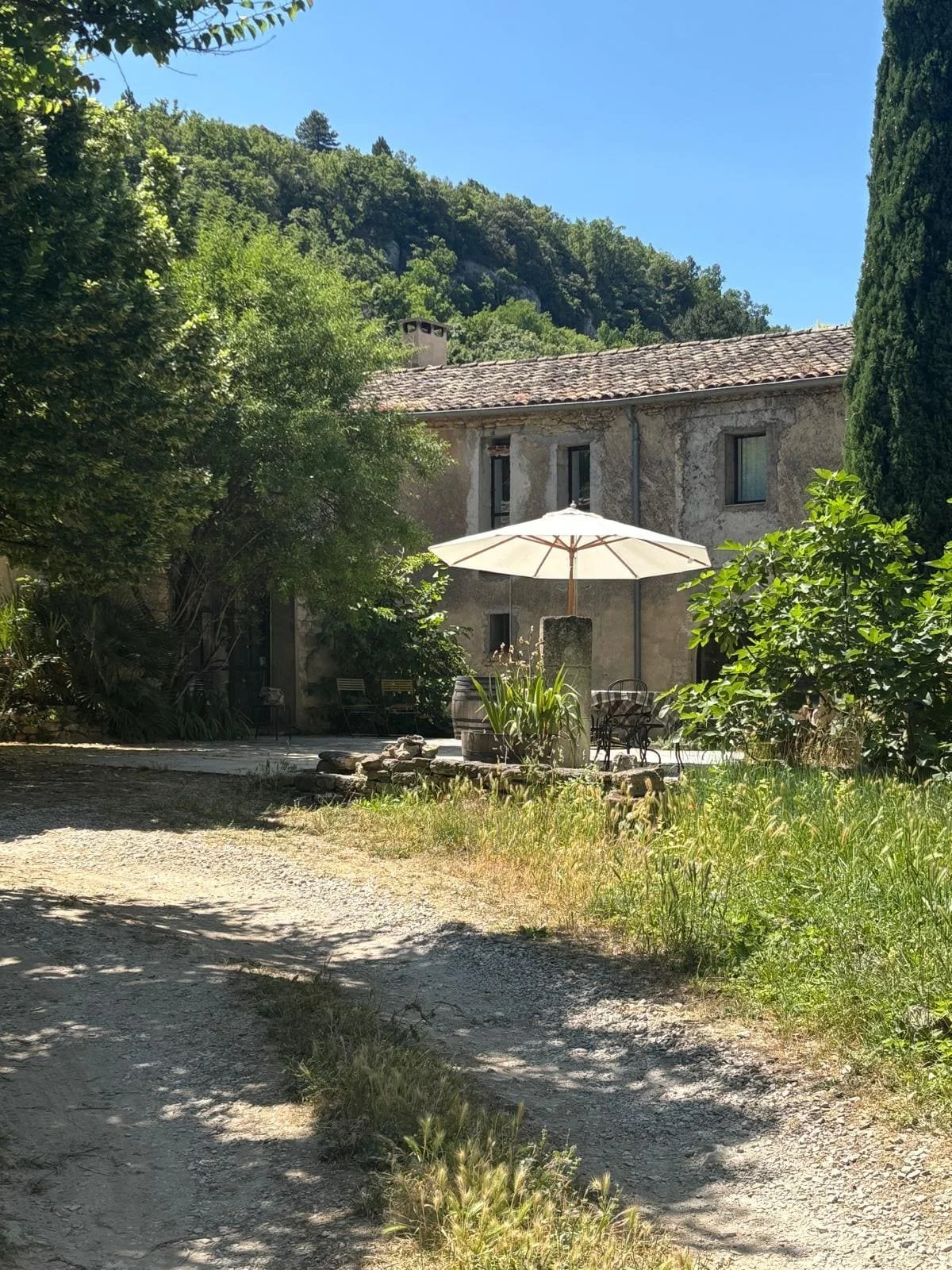 Une maison en pierre avec un toit en tuiles dans un environnement rural, entourée d'arbres et de végétation, sous un ciel bleu. Un parasol blanc et une table avec des chaises sont visibles dans la cour.