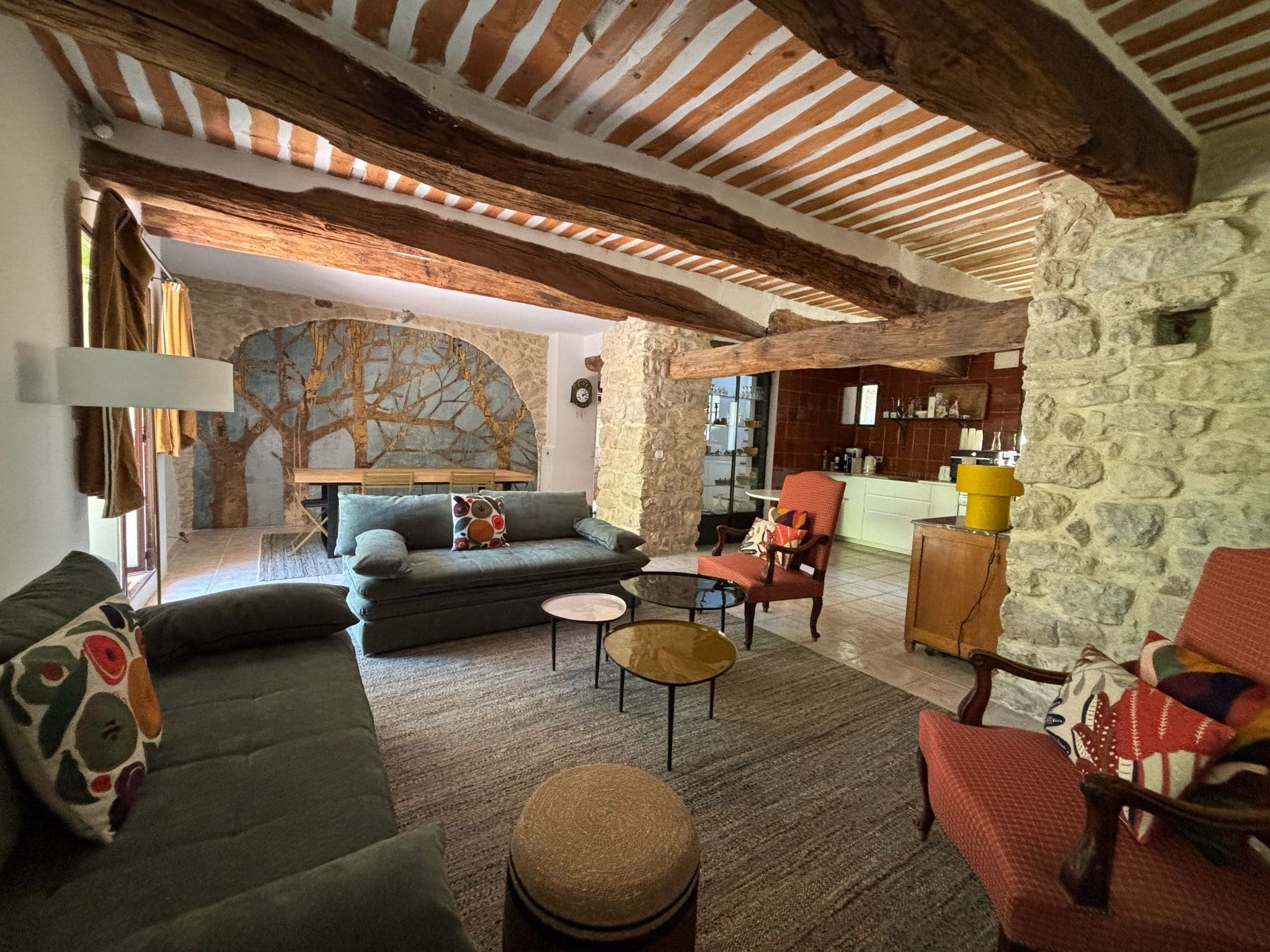 Salon avec deux canapés et deux fauteuils, table basse, mur en pierre, poutres en bois, mural avec arbre peint, cuisine en arrière-plan.