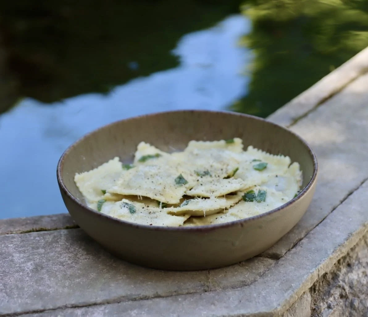 Une assiette de raviolis à la sauce, garnis de morceaux de basilic, placée sur une pierre près d'un cours d'eau calme en plein air.