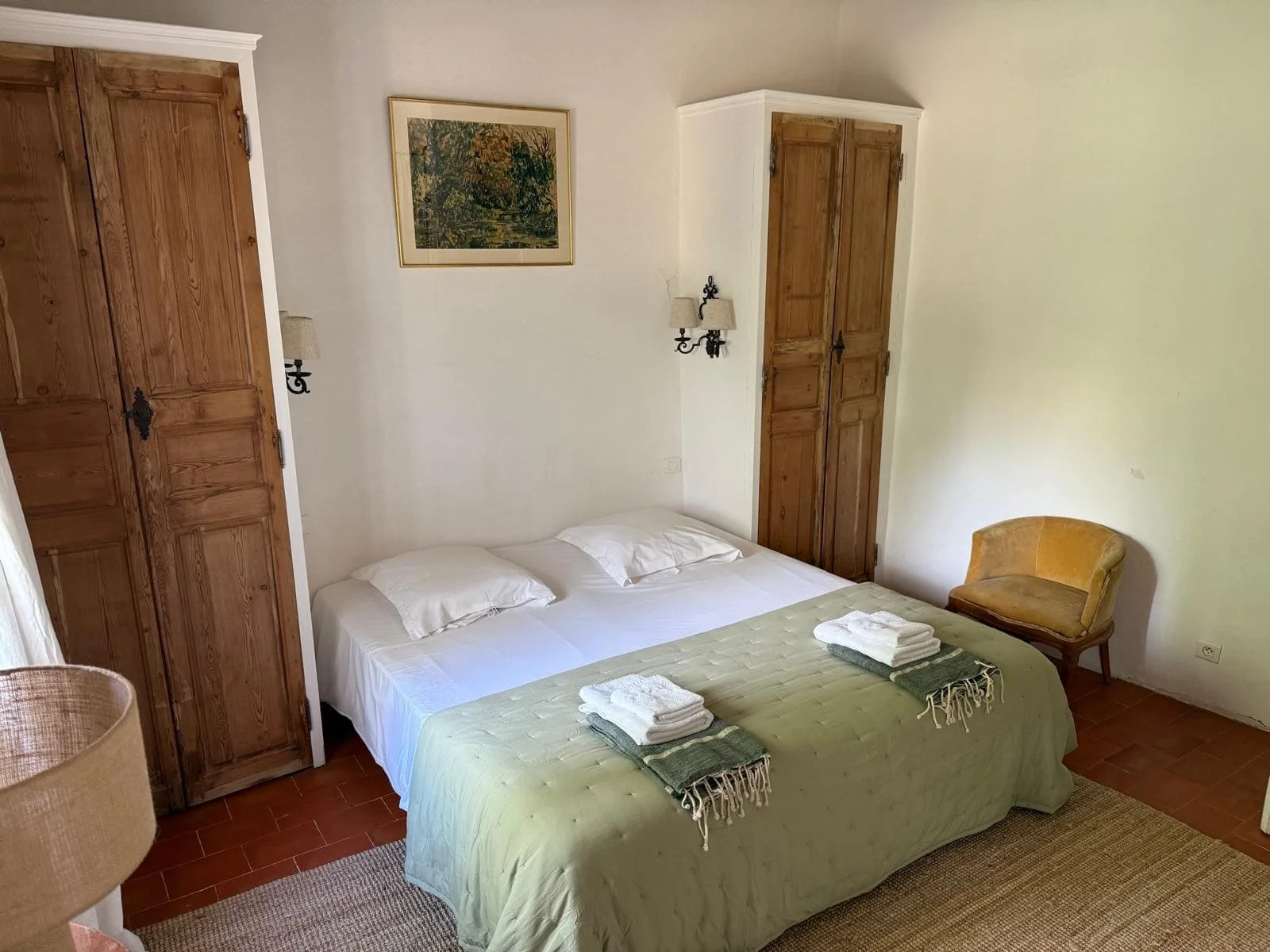 Chambre avec un lit double, deux armoires en bois, une chaise ancienne, une lampe murale, une peinture et des serviettes pliées sur le lit.