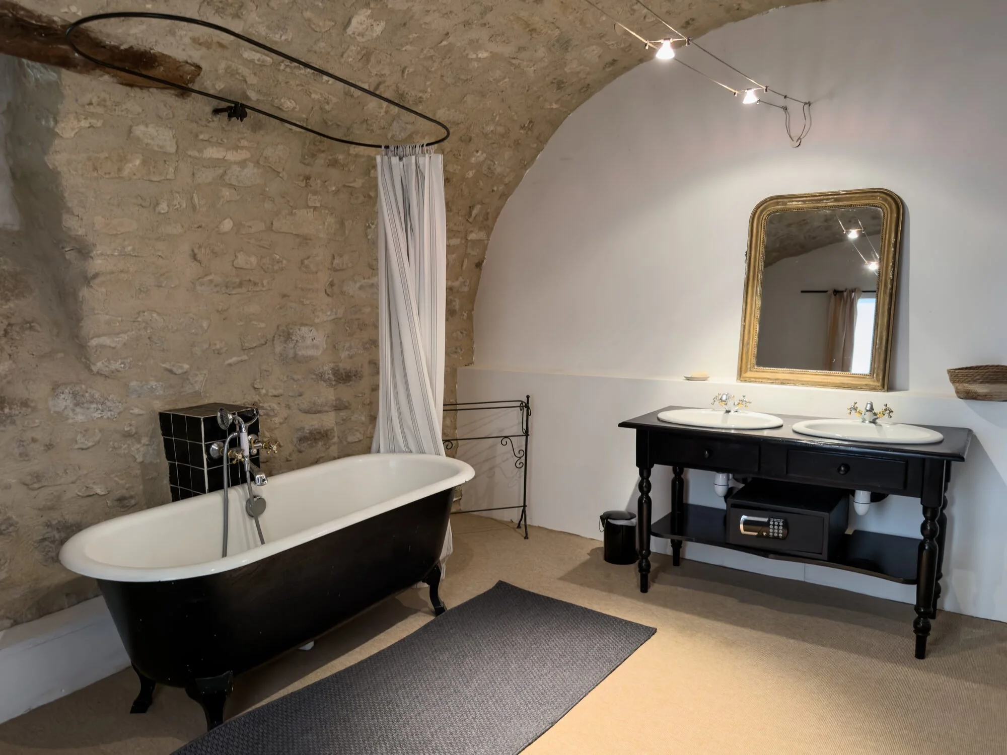 Bathroom with clawfoot bathtub, separated by a white curtain, stone wall, sink with gold mirror, black cabinet, carpeted floor.