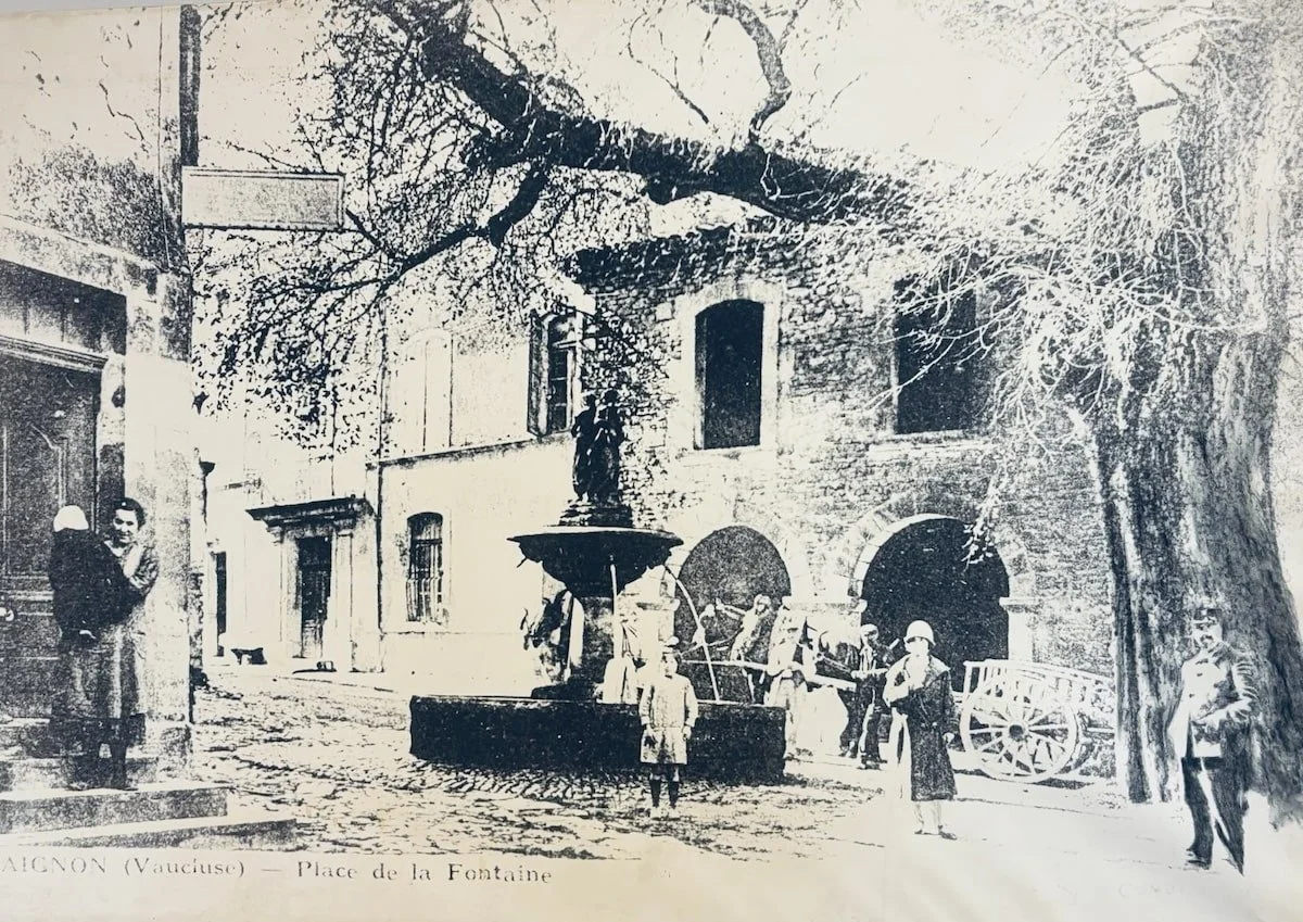 An old photograph showing a square with a central fountain, people standing around it, trees, and a stone building in the background.