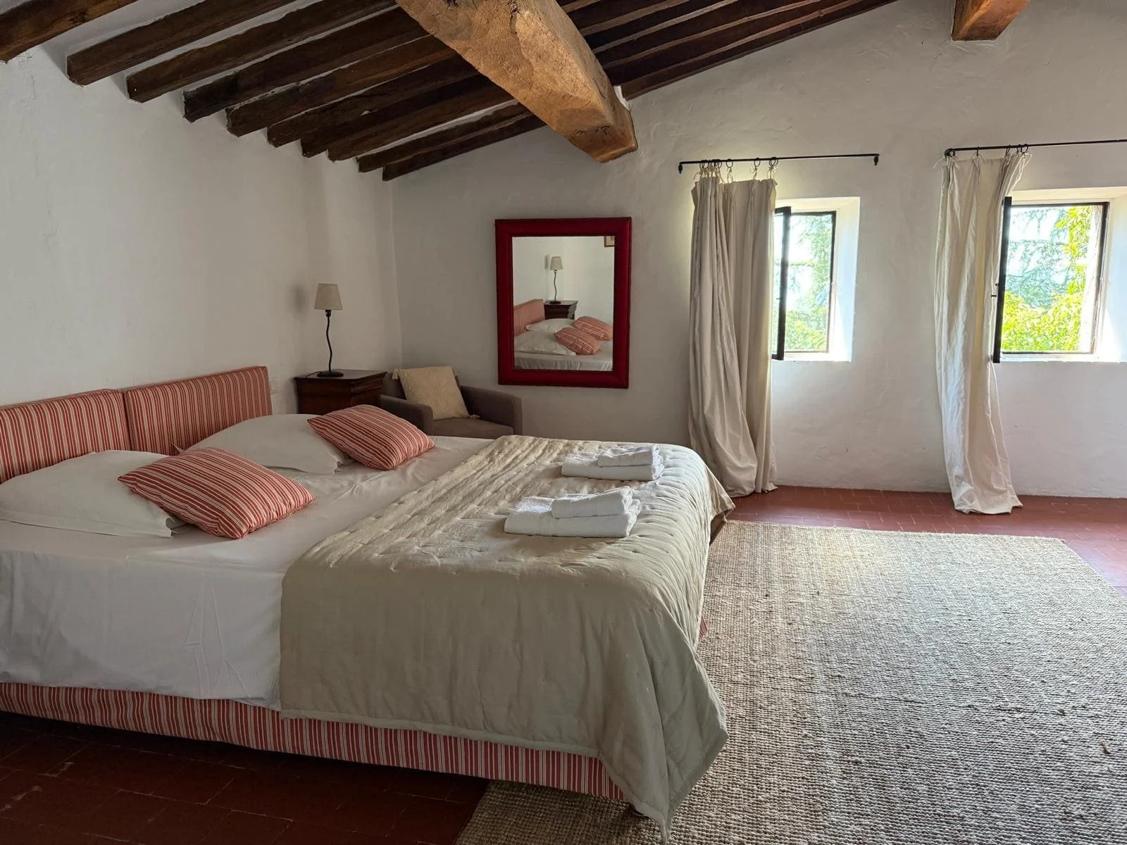 Chambre avec lit double, rayures rouges, deux oreillers rayés rouges et blancs, deux serviettes pliées sur le lit, tapis beige, deux fenêtres ouvertes avec rideaux blancs, miroir avec cadre rouge, lampes murales, murs blancs, plafond en bois.