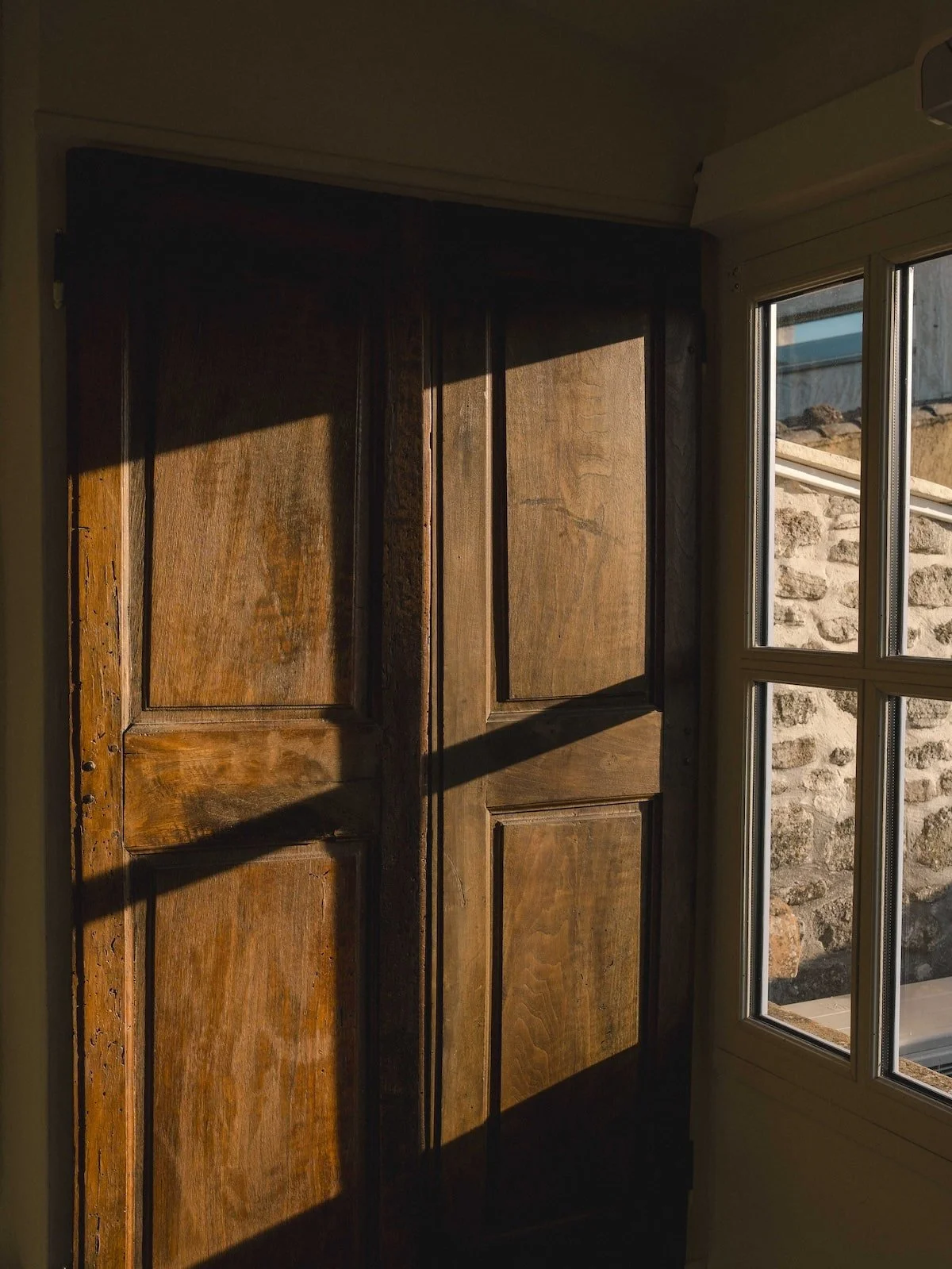 Une porte en bois avec des détails en relief, laissant passer la lumière qui crée des ombres. À côté, une fenêtre avec des vitres, donnant sur un mur en pierre.