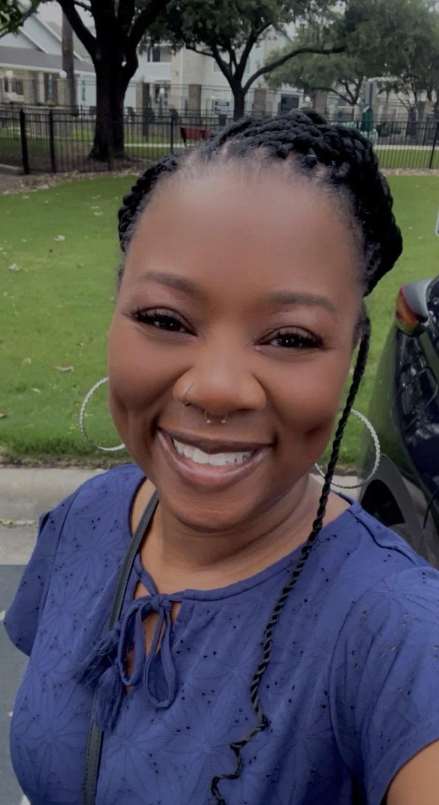 A smiling woman with braided hair, large hoop earrings, and a nose ring taking a selfie outdoors in a park with green grass, trees, and residential buildings in the background.