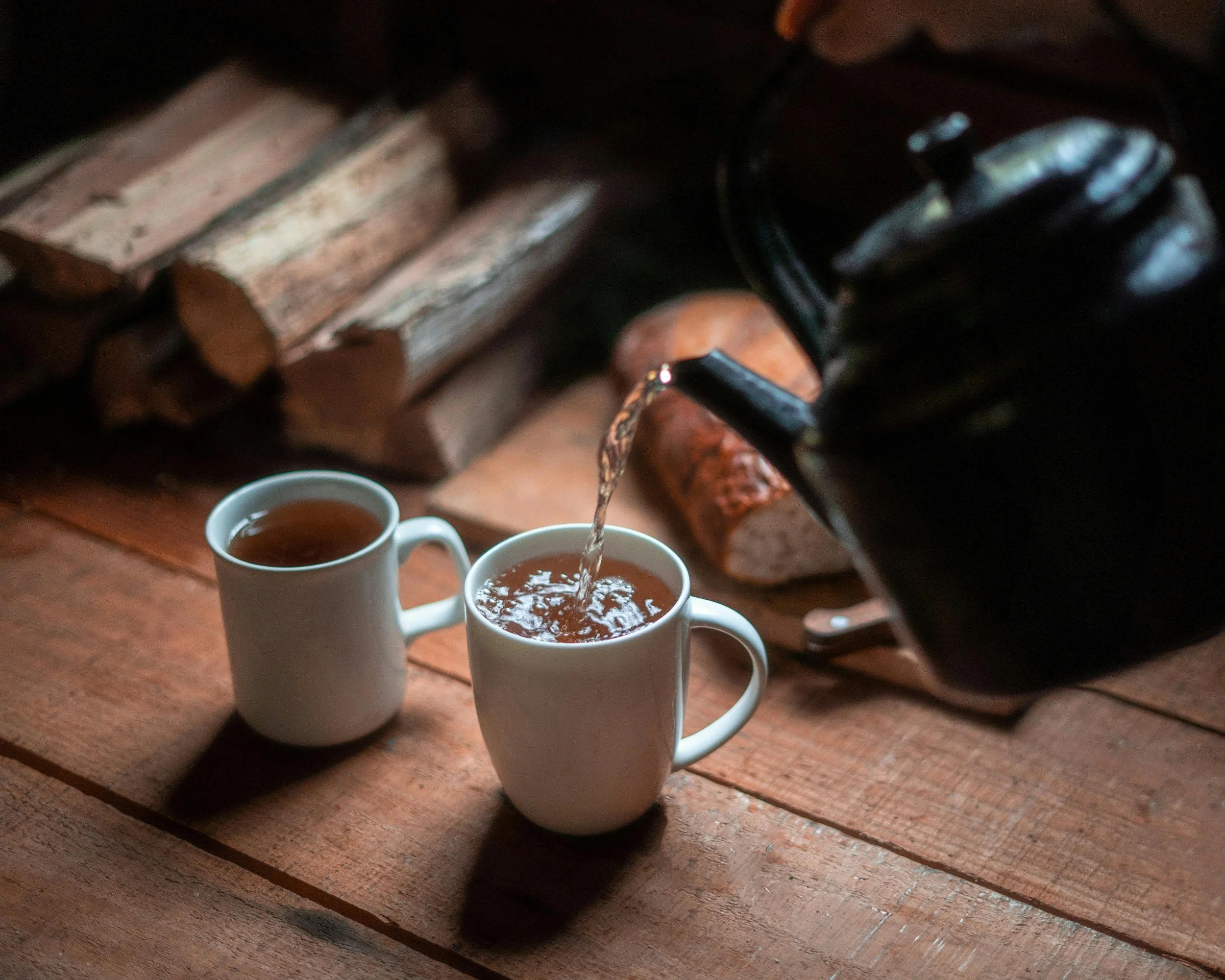 A black teapot pouring hot tea into a white mug, with a second mug nearby, on a wooden table with firewood in the background.