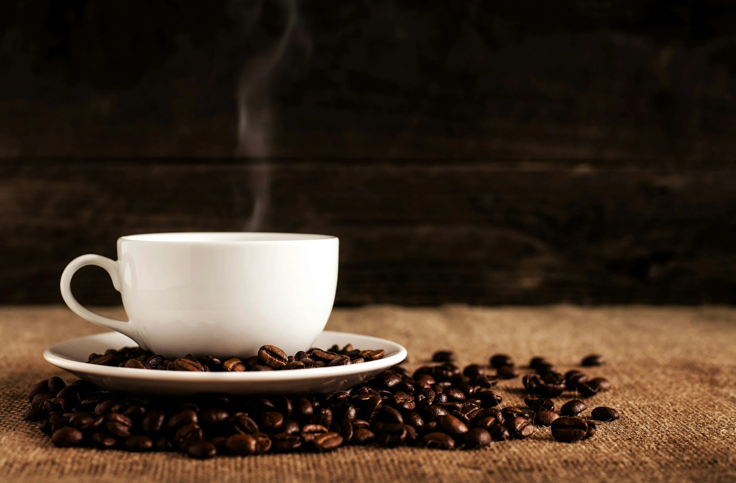 A white coffee cup on a saucer surrounded by roasted coffee beans on a brown textured surface with a dark wooden background, steam rising from the cup.