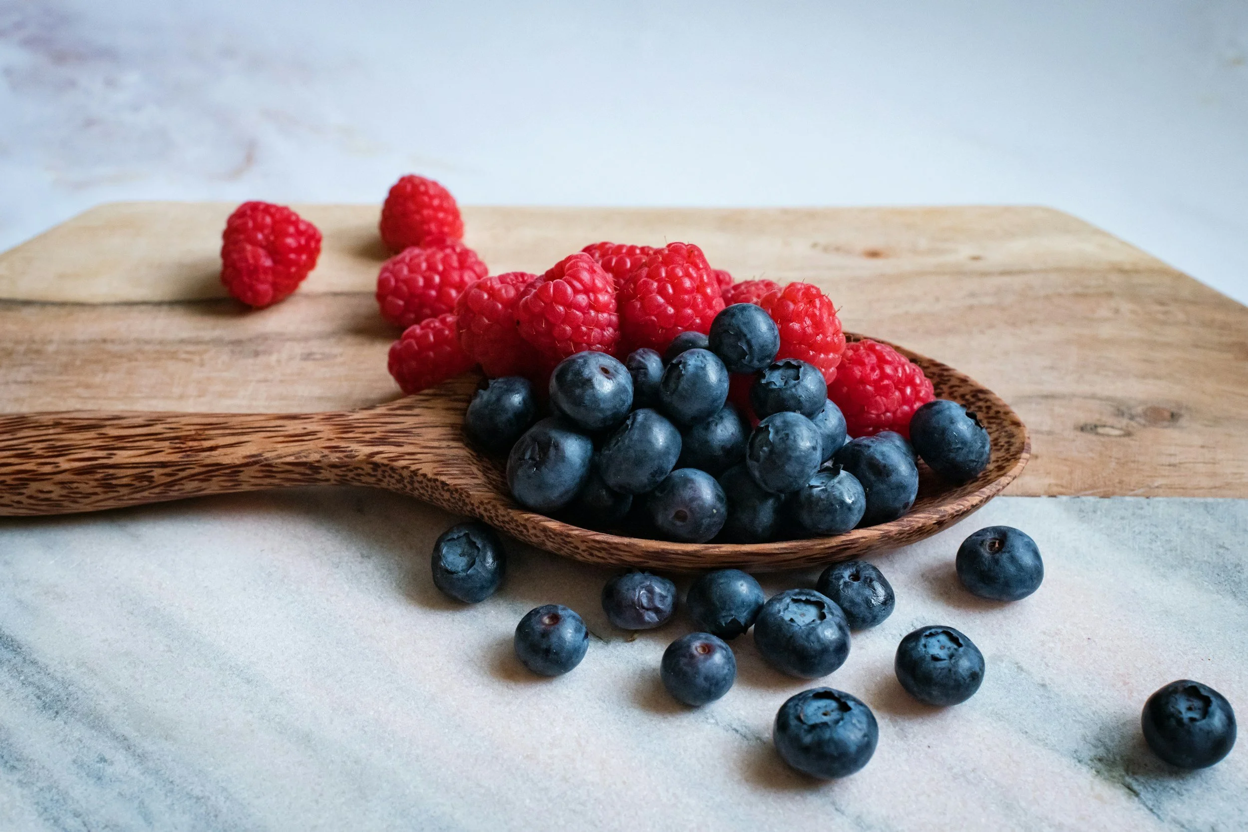 Fresh red raspberries and blueberries on a wooden spoon and scattered on a surface with a wooden cutting board in the background.