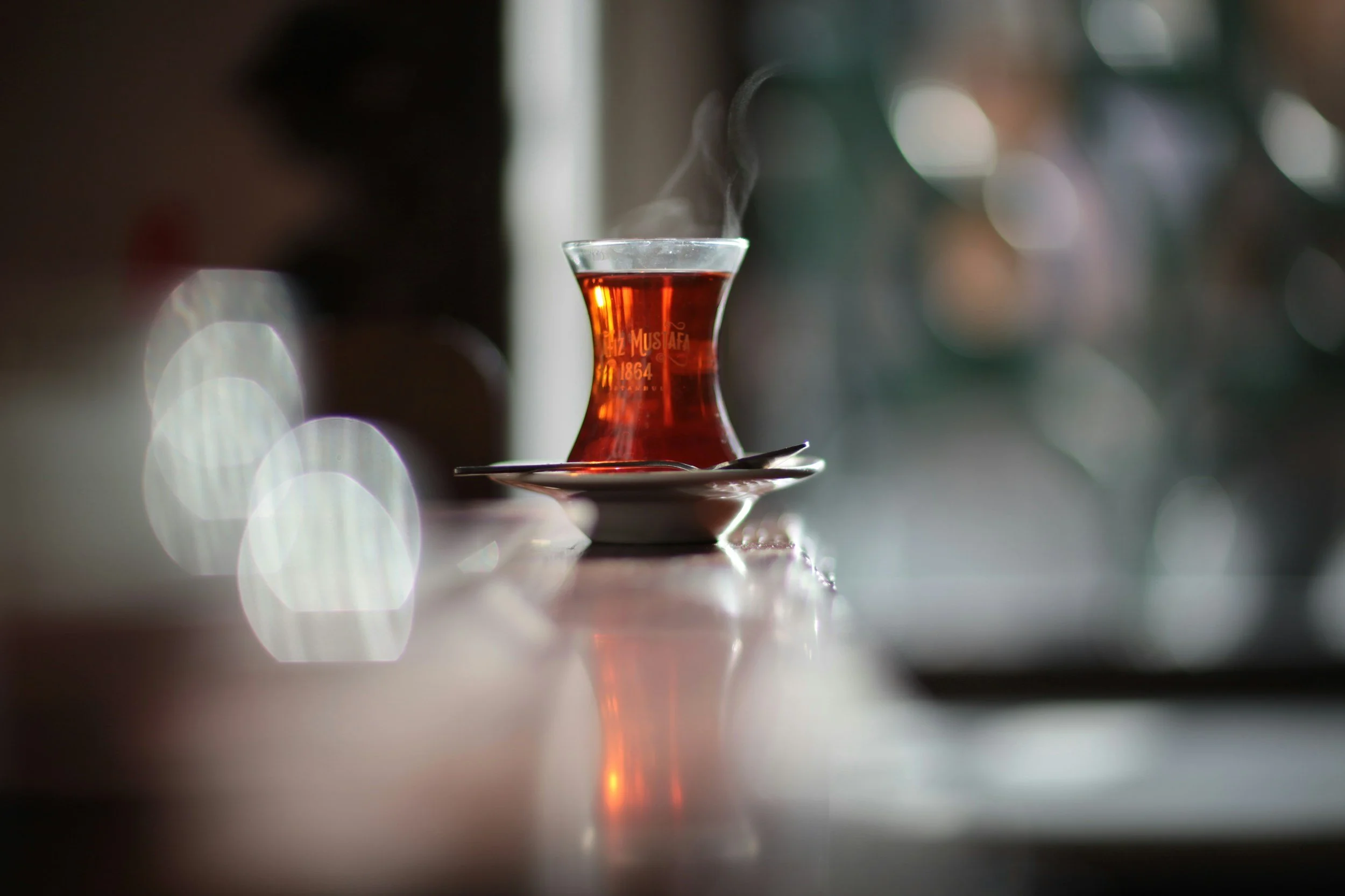 A glass of hot tea with steam rising, placed on a saucer with a spoon, on a reflective surface in a dimly lit setting.