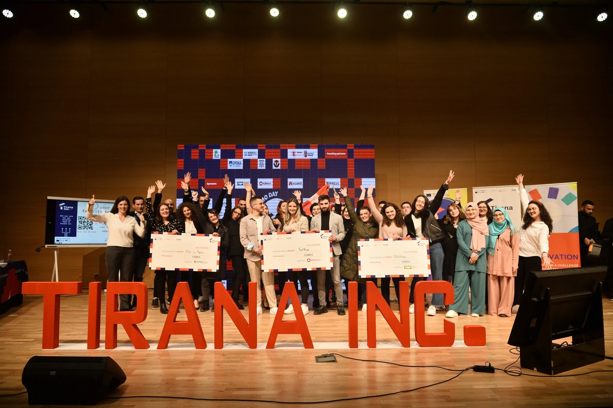 Group of people on stage celebrating, holding large checks, in front of a large Chromebook-like screen and red 3D letters spelling 'TIRANA INC.' at an indoor event.