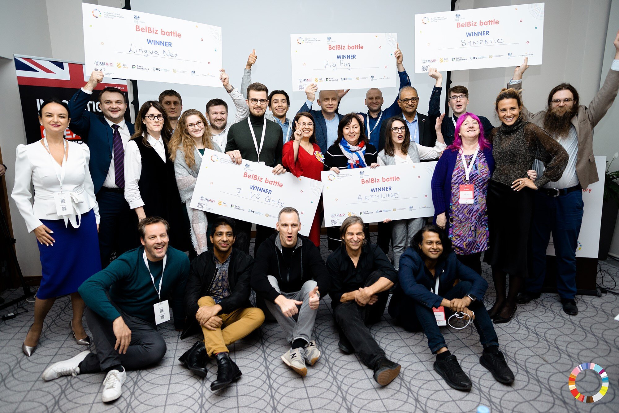 Group of diverse people celebrating with oversized checks at an event, holding up signs with winners' names, in a conference room with a gray wall and patterned carpet.