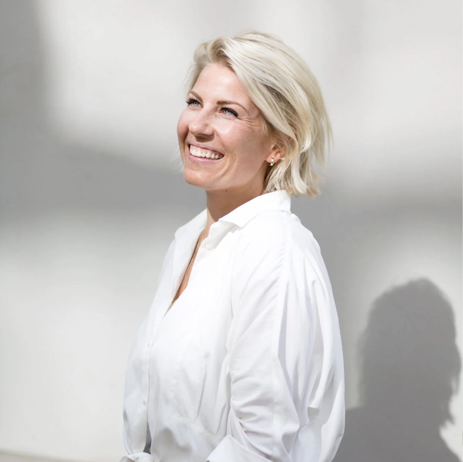 A woman with short blonde hair smiling and wearing a white shirt against a plain background.