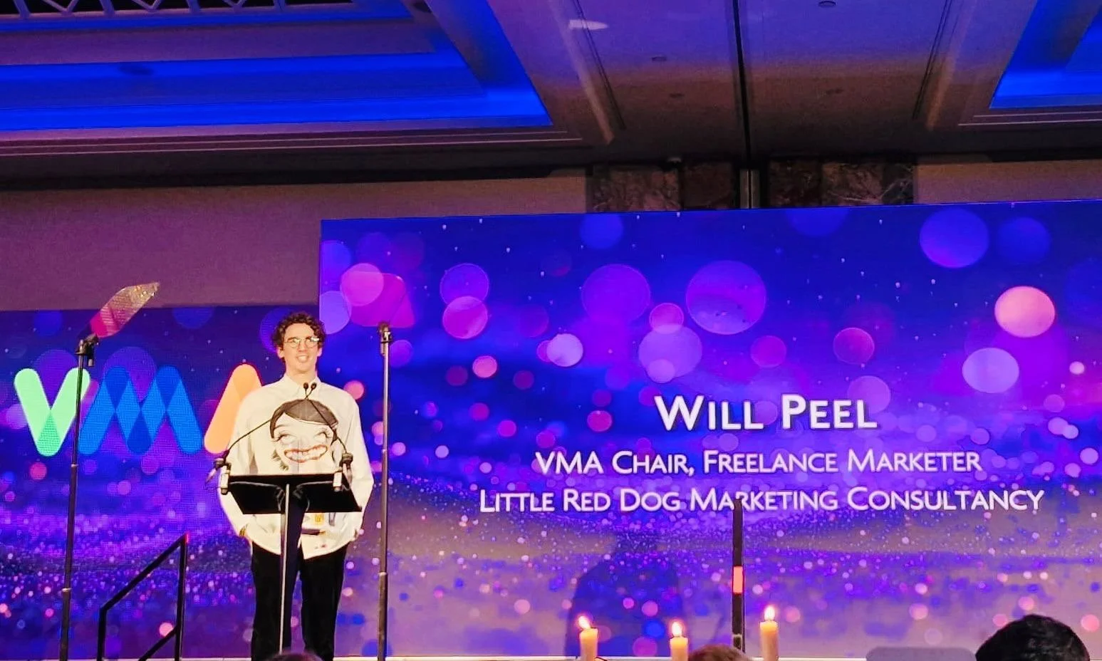 Will Peel stands on stage with a microphone, giving a speech at a conference, with a large screen behind him displaying his name, title, and company, and a colorful background.