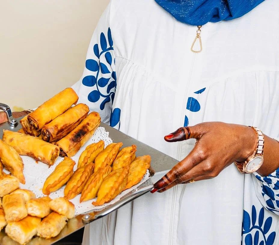 Person holding a tray of assorted fried snacks including spring rolls and other pastries, wearing a white and blue outfit with a blue headscarf.