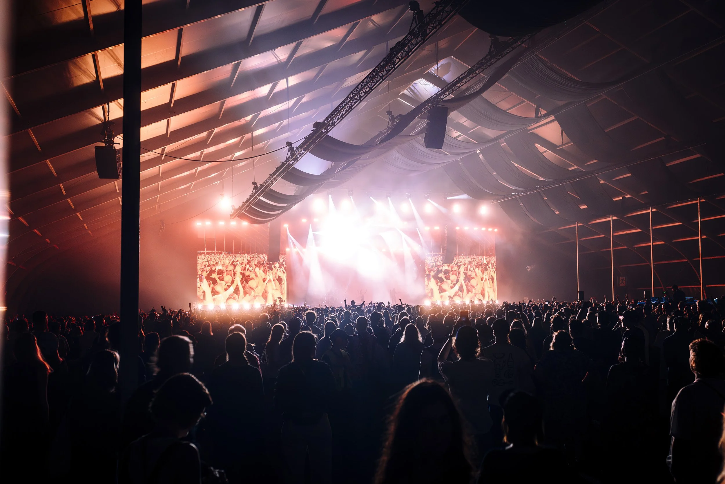 Suuri yleisö konserttitilassa, jossa on valo- ja videoprojisointeja taustalla, ja ihmiset nauttivat musiikista.