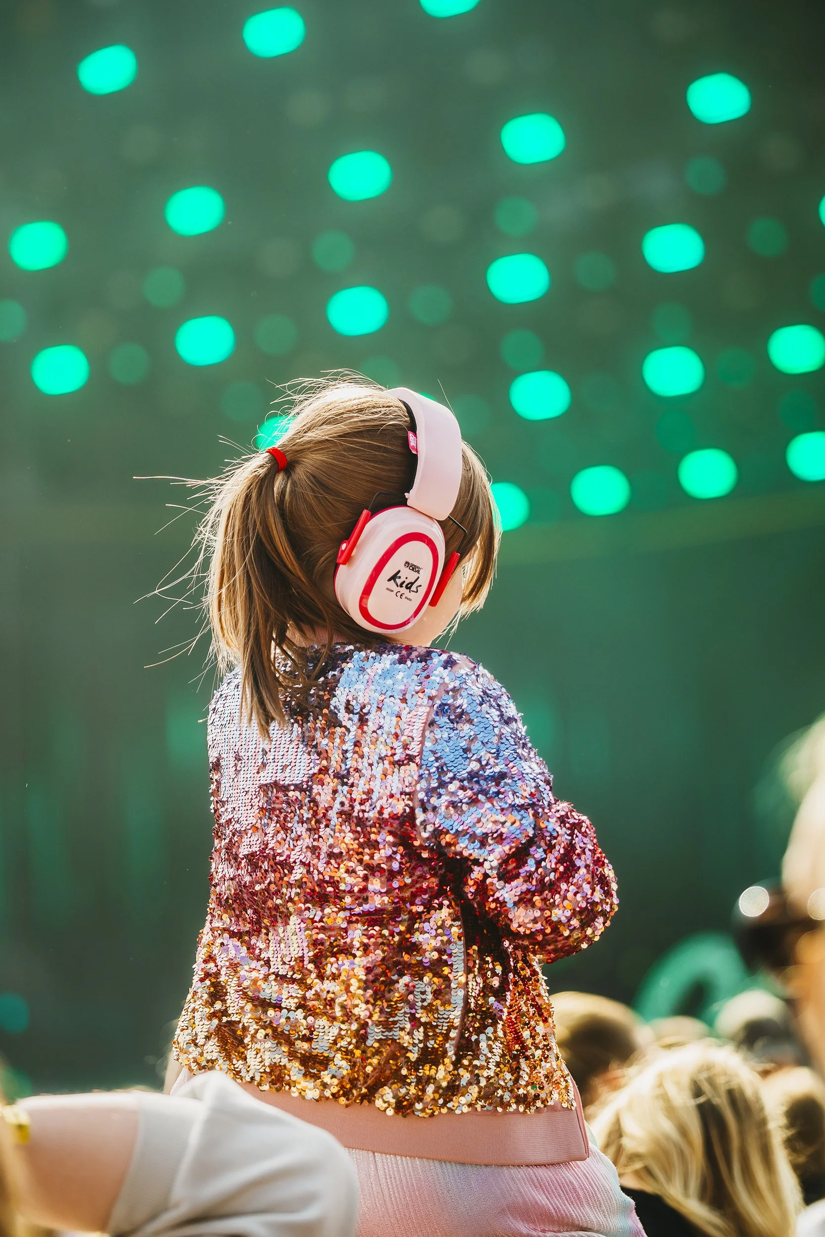 Pieni tyttö kuuntelee musiikkia lasten kuulosuojaimet päässä. Taustalla vihreitä valoja.