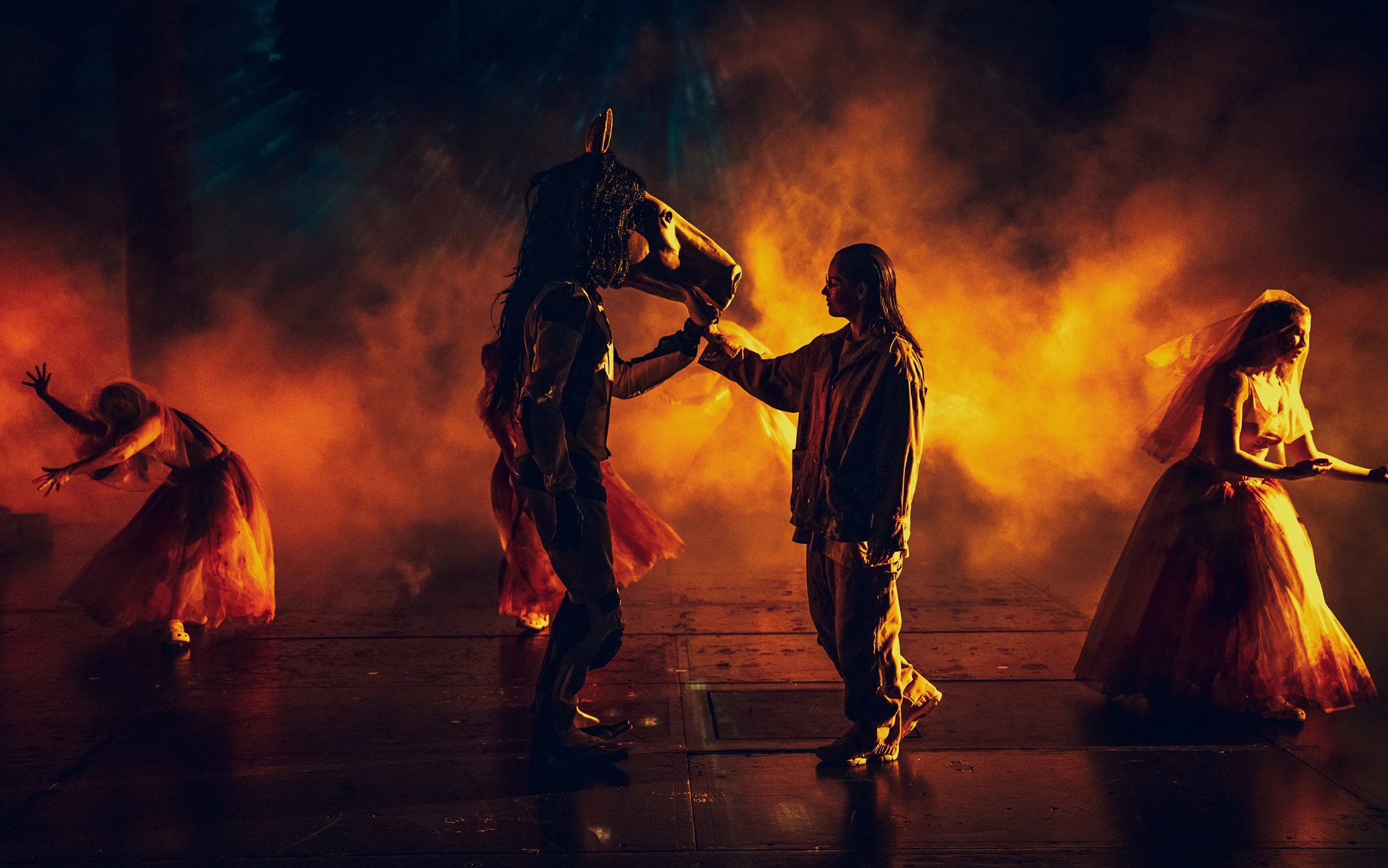 Tanssijoita lavalla, taustalla savua ja tulta, keskellä henkilö maskin ja vestin kanssa, toinen naista heiluttaen käsillään.