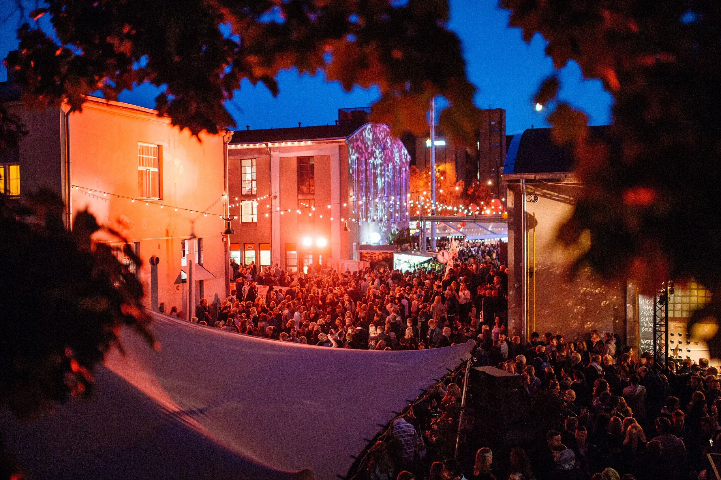 Ihmisiä iltahämärässä festivaaleilla, jossa on paljon tunnelmallisia valoja ja telttoja, ja taustalla rakennuksia ja puiden oksia.