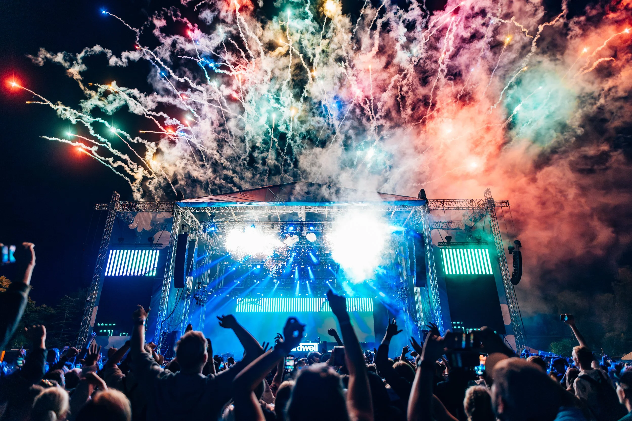 Yleisö on kokoontunut konserttiin tai festivaaliin, jossa lavalla on paljon valoja ja äänentoistolaitteita. Taivaalla raketit ja ilotulitukset räjähtävät värikkäinä sateina yläilmakehässä.
