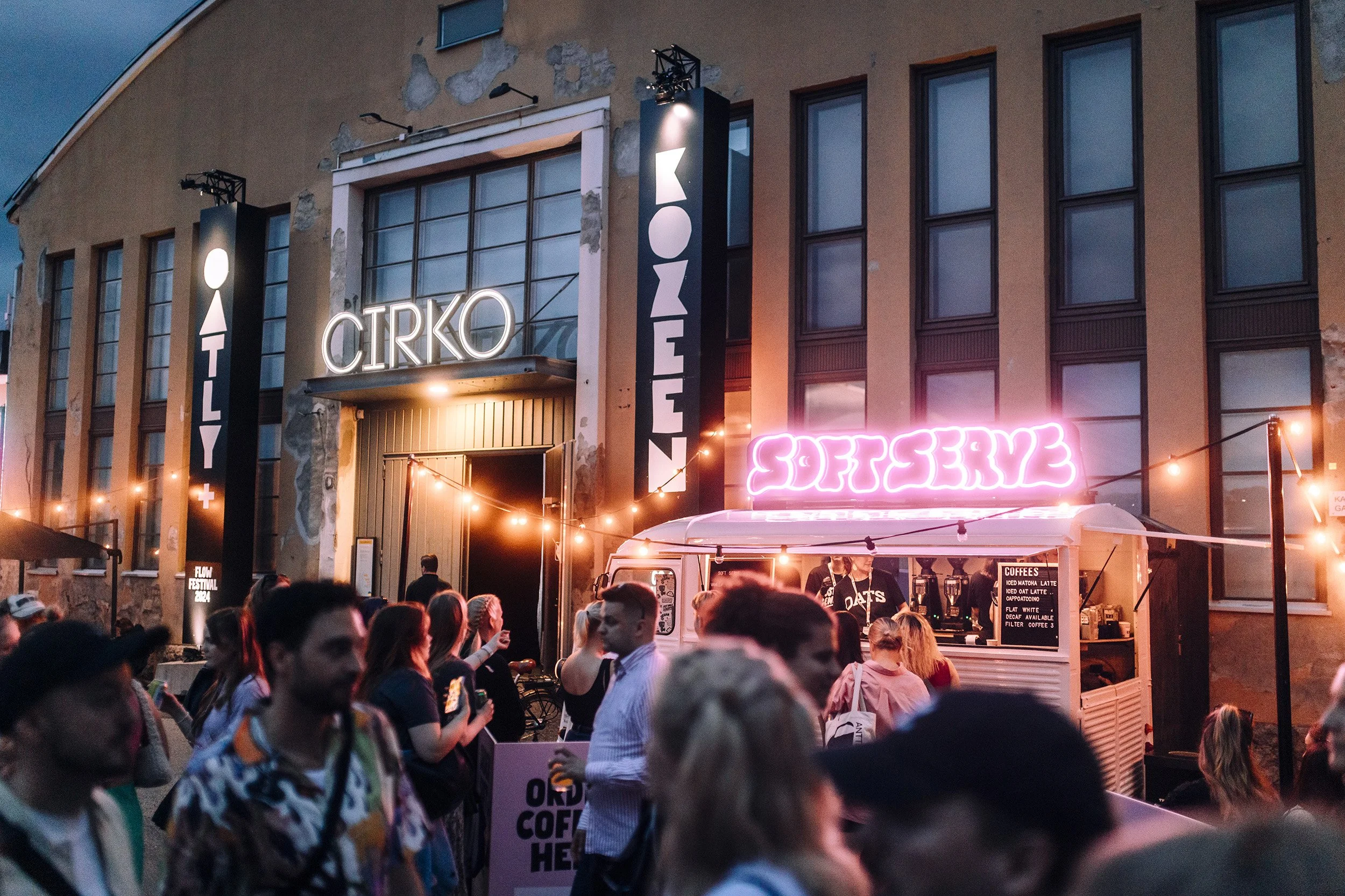 Kuva ulkopuolisesta tapahtumasta, jossa ihmiset odottavat ja nauttivat kahvista ja esityksistä ulkona, teatteritalon edessä, jossa on neonkyltit 'COFFEE' ja 'CIRKO', myyntikoju ja koristevalot