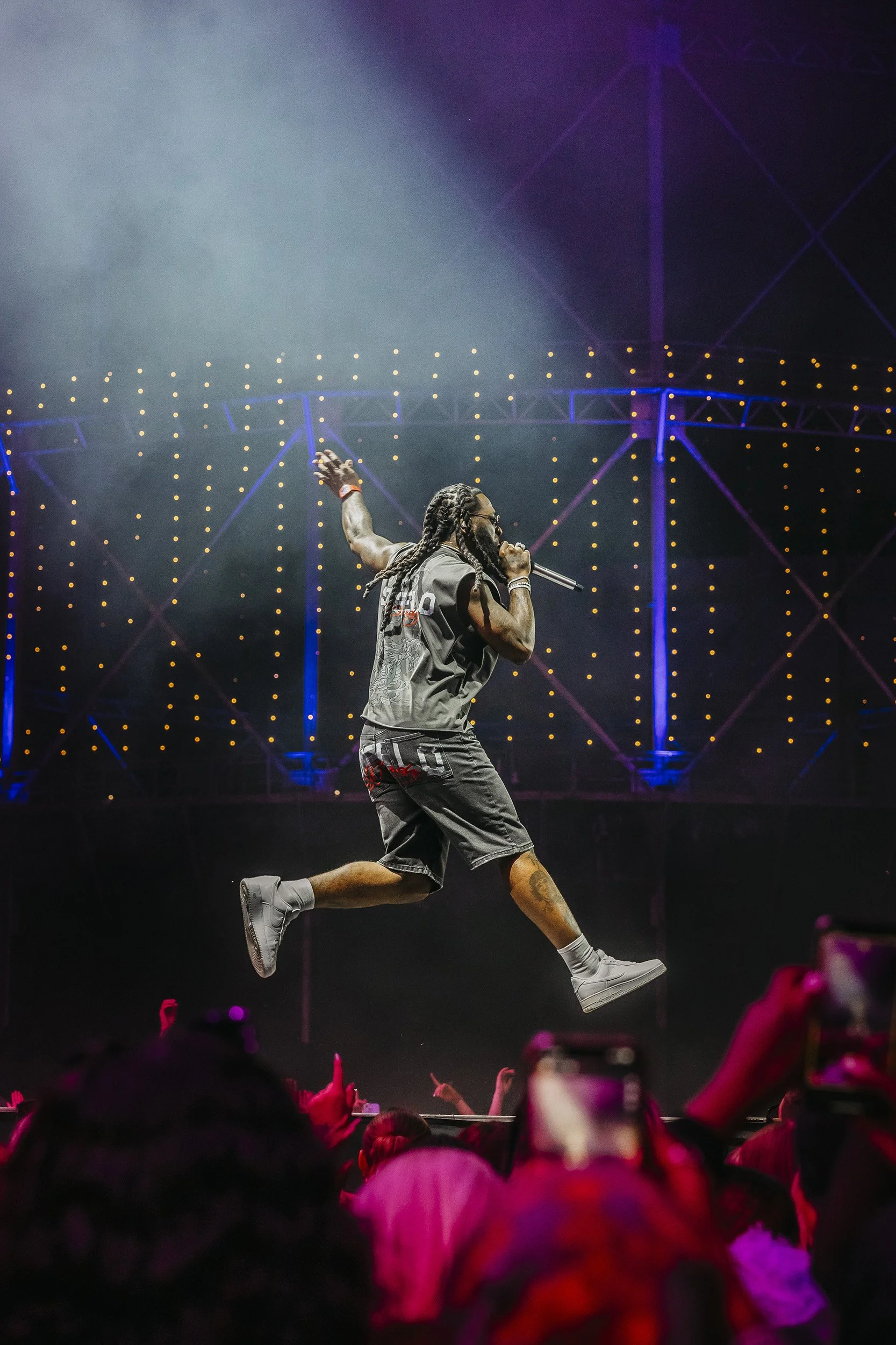 Rap artisti esiintyy lavalla, hypäten ilmaan ja laulamalla mikrofoniin, taustalla valoja ja rakenteita.