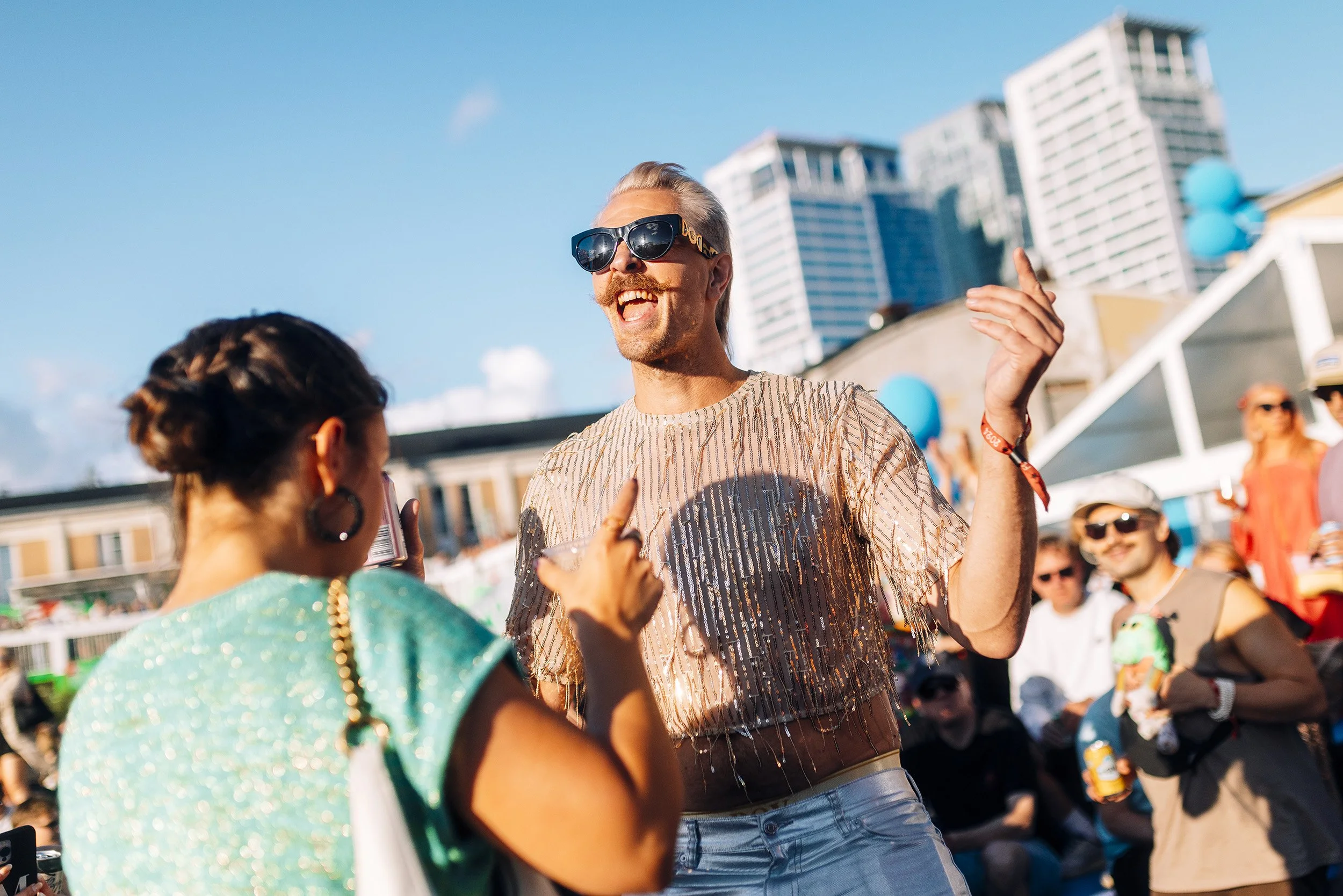 Ihmiset nauttimassa tapahtumasta ulkona auringonpaisteessa, ohutta vaatetta ja aurinkolaseja, kaupunkimaisemalla taustalla.