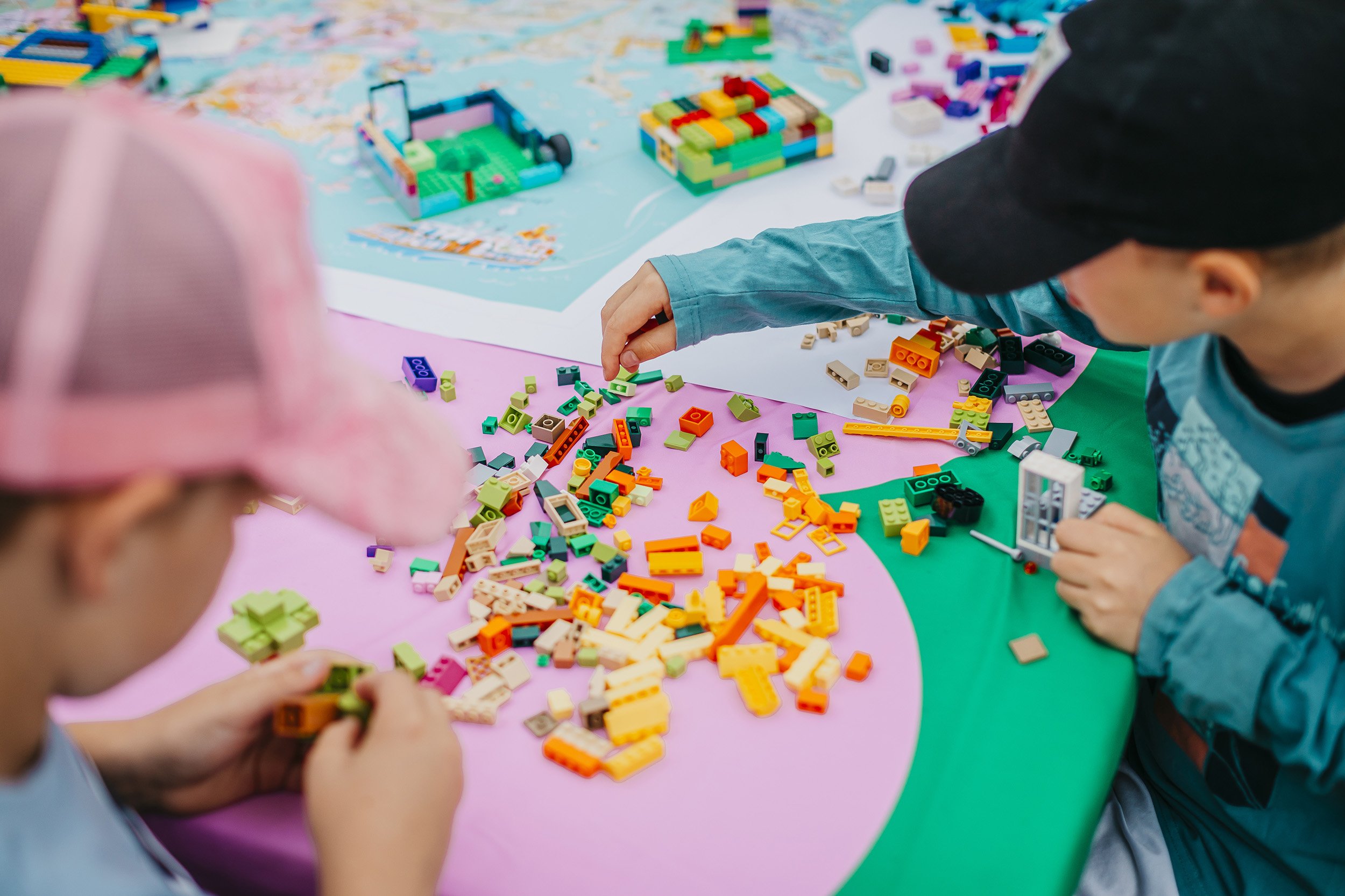 Kaksi lasta rakentavat lego-palikoista värikkäitä rakennelmia pöydällä, taustalla suurempi lego-rakennelma.