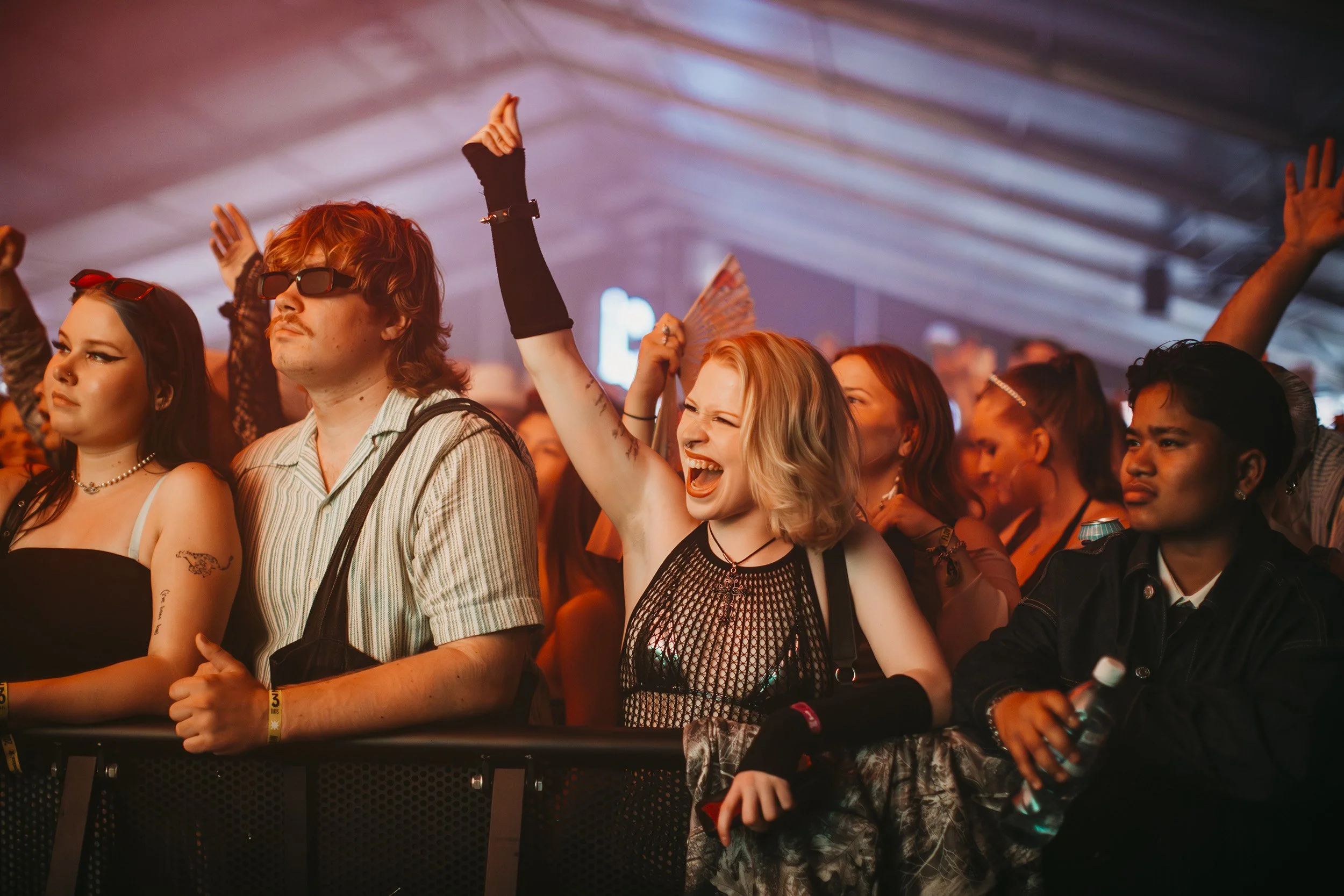 Yleisö konserttilavalla, iloisia ihmisiä, yksi nostaa kättään ylös, taustalla valot ja kattorakenne.