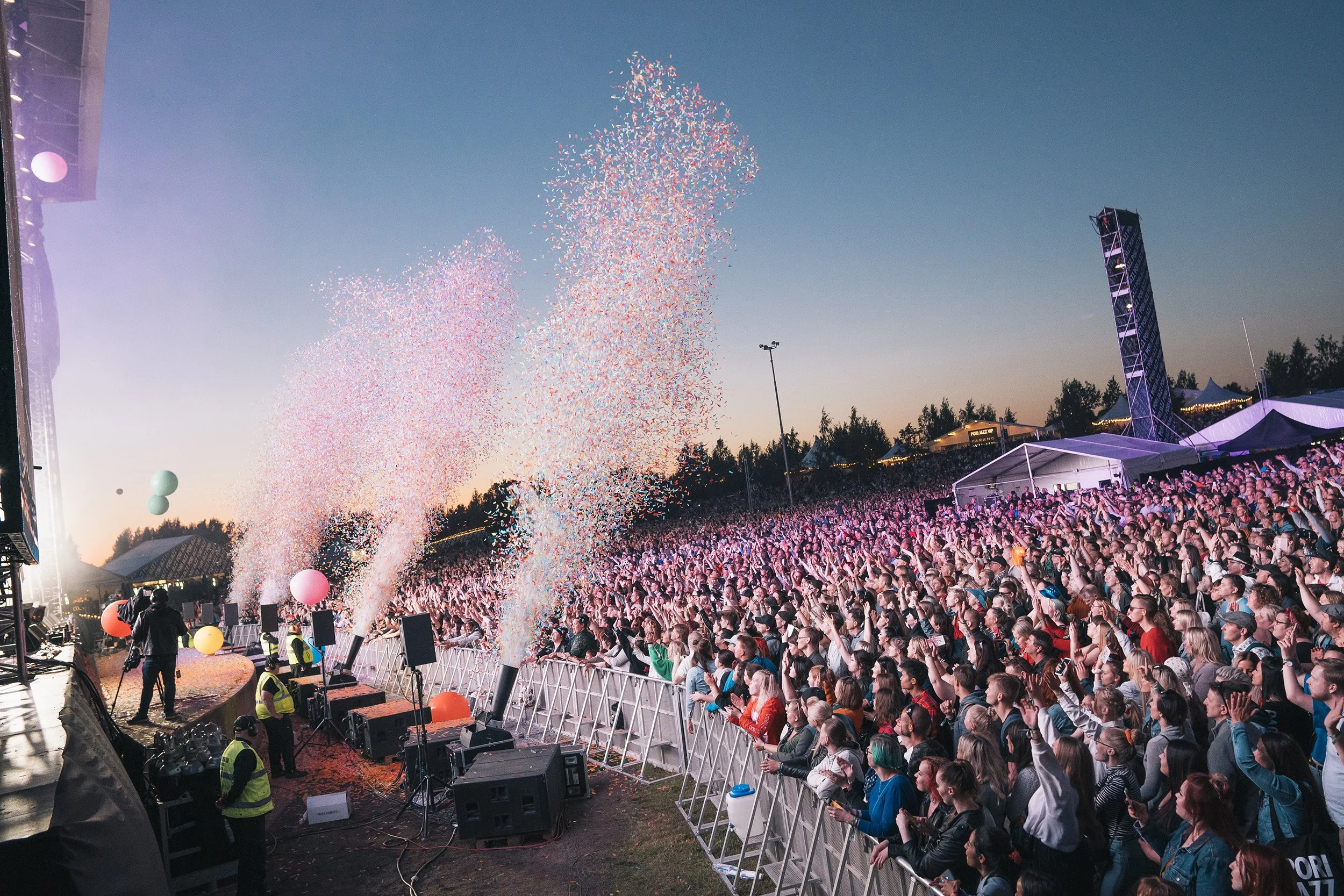 Suuri konserttilava ja väkijoukko nauttimassa tapahtumasta illan hämärtyessä. Ilmassa ilotulitteiden ja värikkäiden konfettien säteilyä.