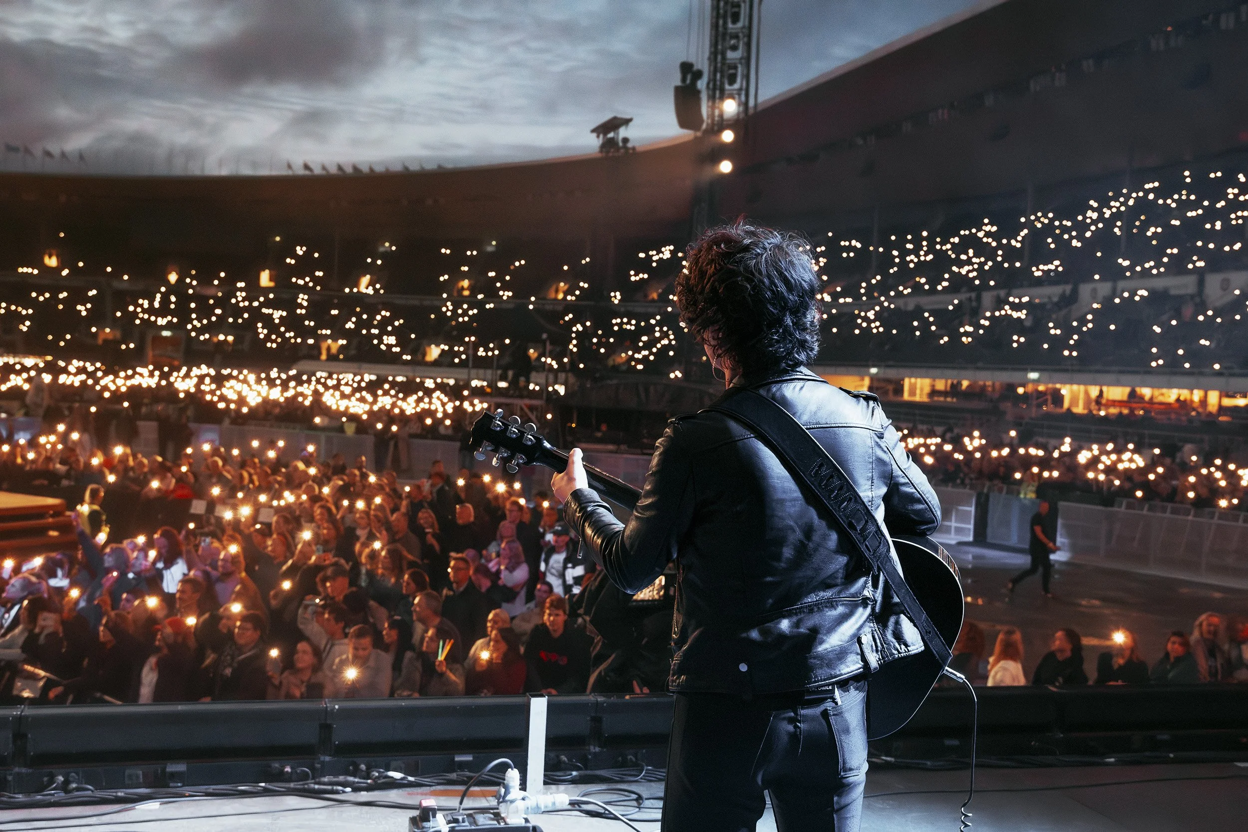 Brittipop tähti esiintymässä lavalla suurella satdionilla, taustalla yleisö pimeässä, täynnä kännykän taskulamppujen luomia valoja.