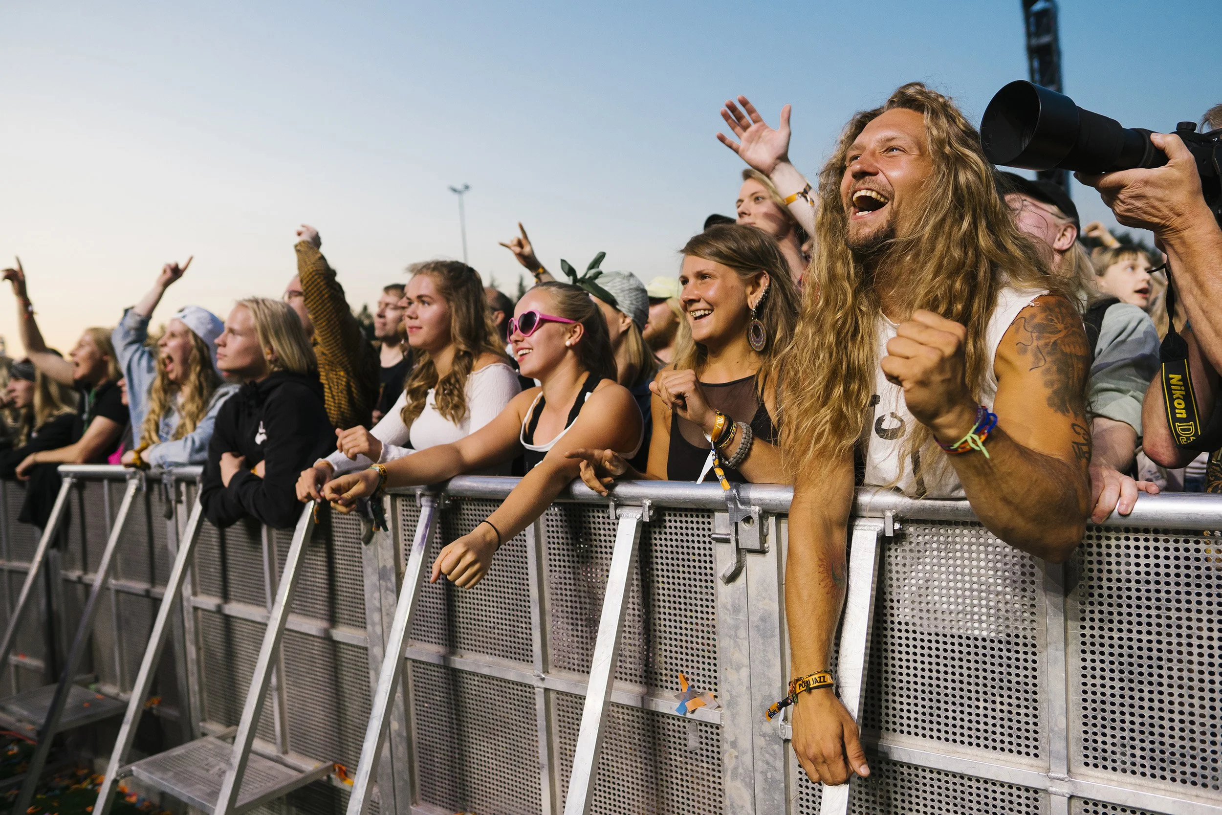 Eturivin yleisö innostuneena musiikkitapahtumassa, ihmiset seisovat metalliaidan takana, osa nauraa ja tanssii, kädet nousevat iloisesti ilmaan, taustalla kirkas ilta-taivas.