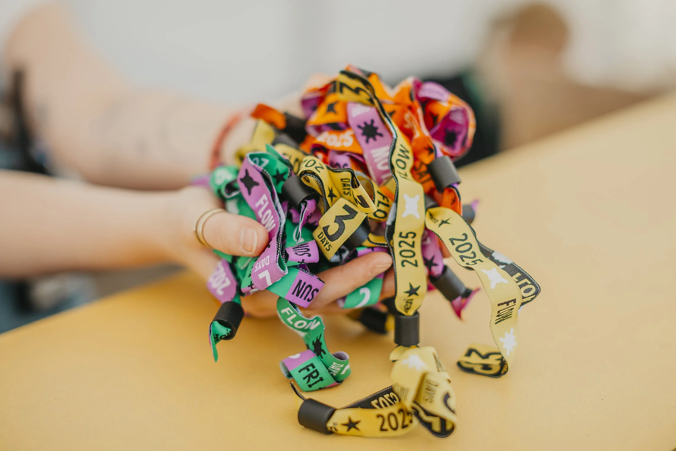 Kuvassa käsi, jossa on kultainen sormus, pitää värikkäitä rannekoruja, joissa on tekstejä kuten "2025", "FLOW FESTIVAL" ja "DAY 3".