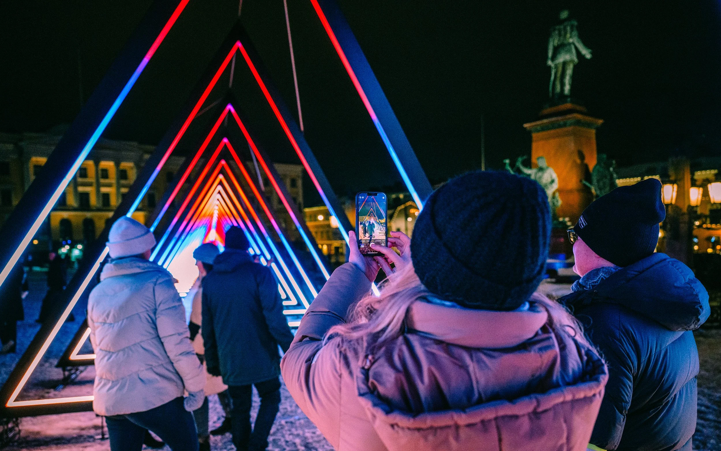 Ihmiset ottavat valokuvia neon-valaistuista kolmioista yöllä.