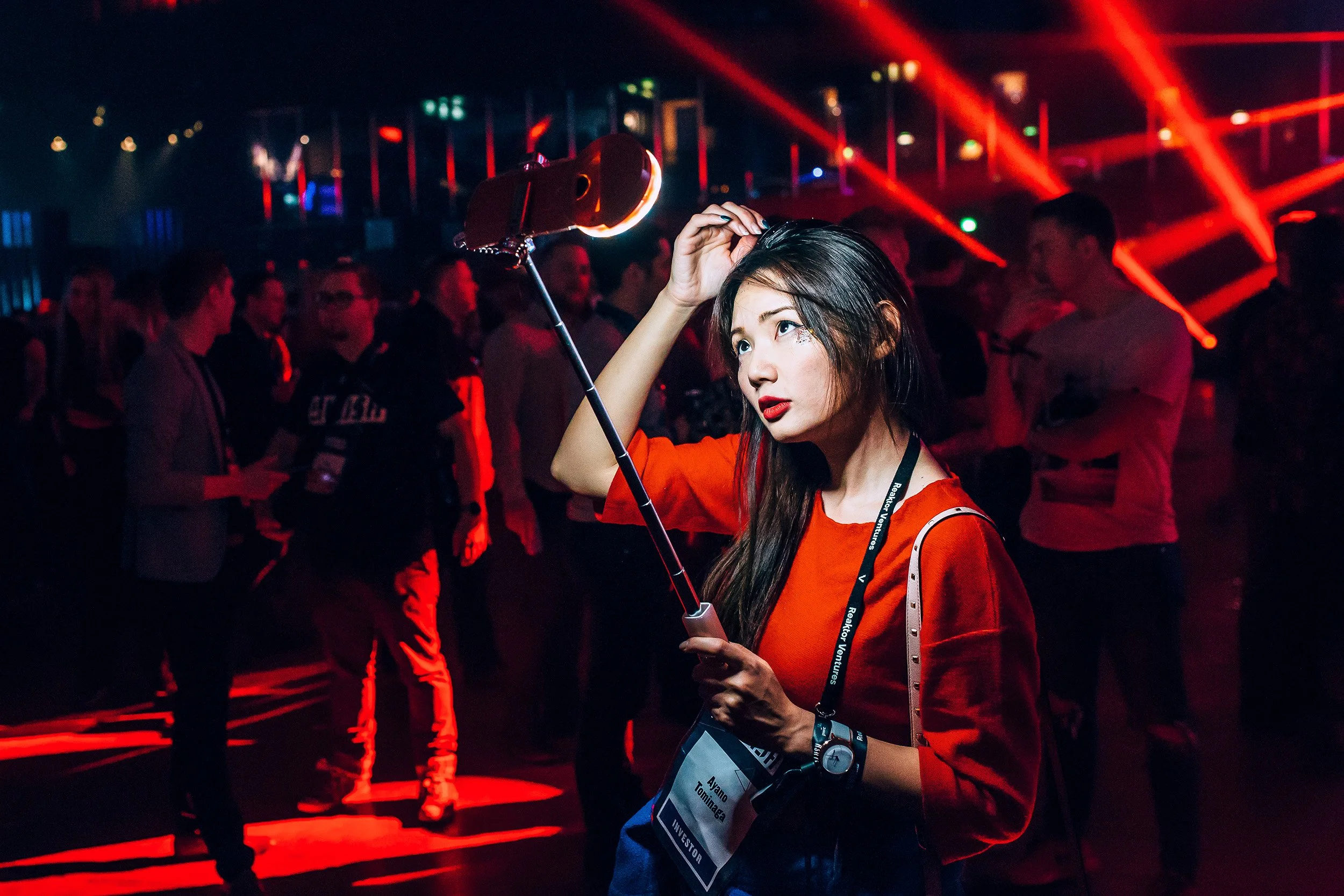 Nuori nainen hämyisässä tapahtumassa, pidellen selfie-valoa, taustalla monia ihmisiä ja värikkäitä valoefektejä.