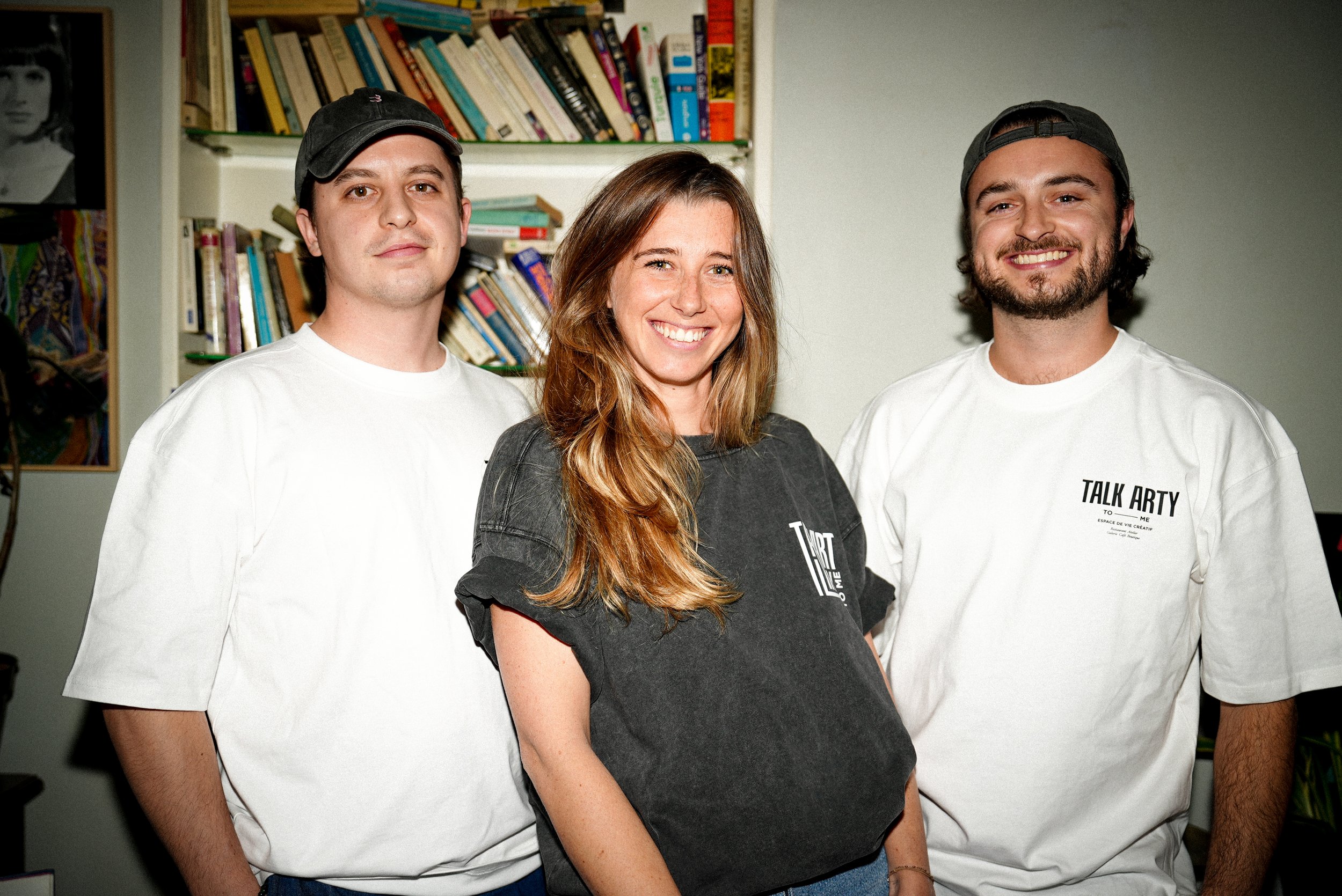 Trois jeunes adultes souriants posant côte à côte dans une pièce, avec une étagère remplie de livres en arrière-plan. Deux hommes portent un t-shirt blanc avec l'inscription 'TALK ARTY', la femme porte un t-shirt gris avec le logo talk arty to me.