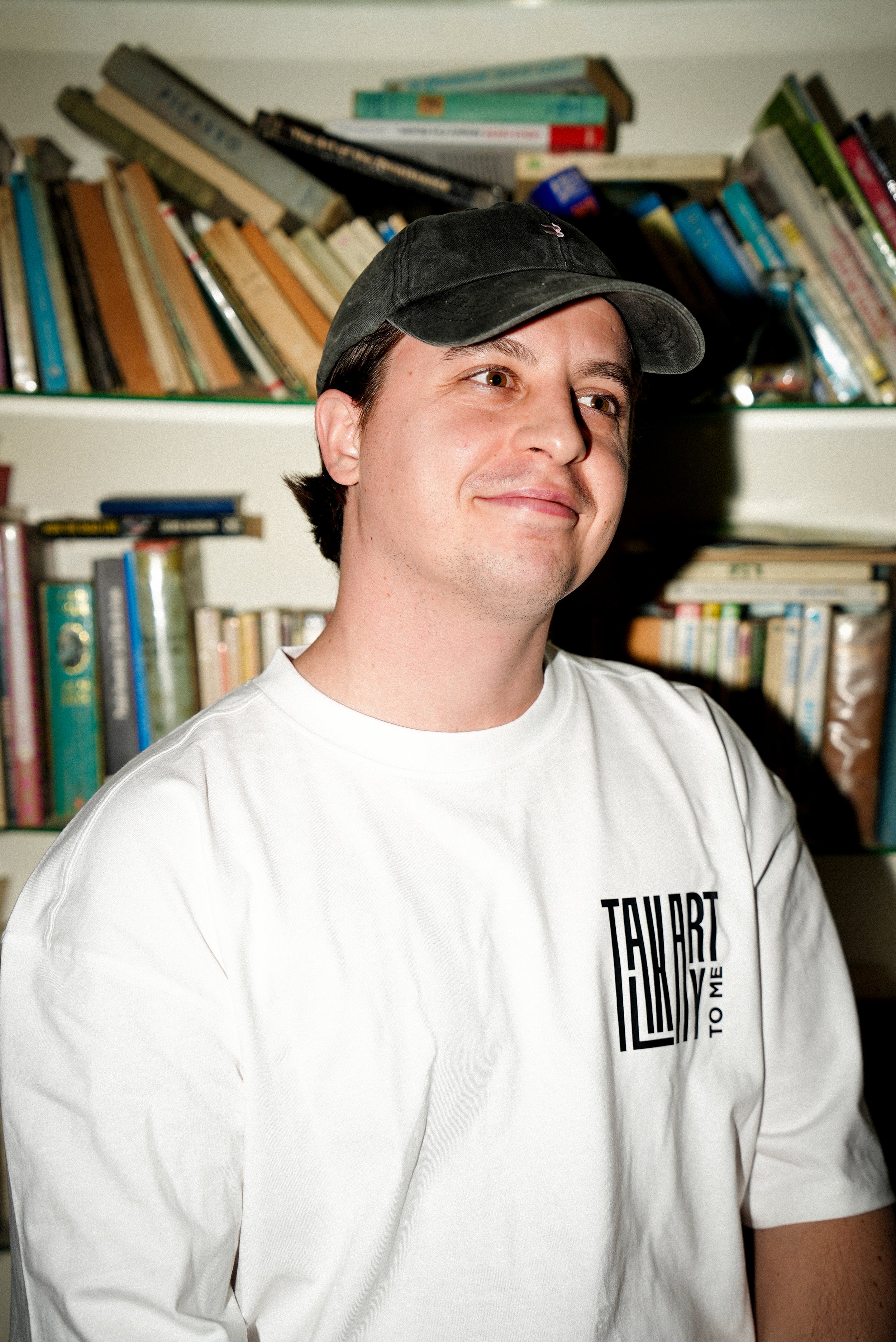 Jeune homme souriant portant une casquette noire et un t-shirt blanc avec inscription, assis devant une étagère remplie de livres.