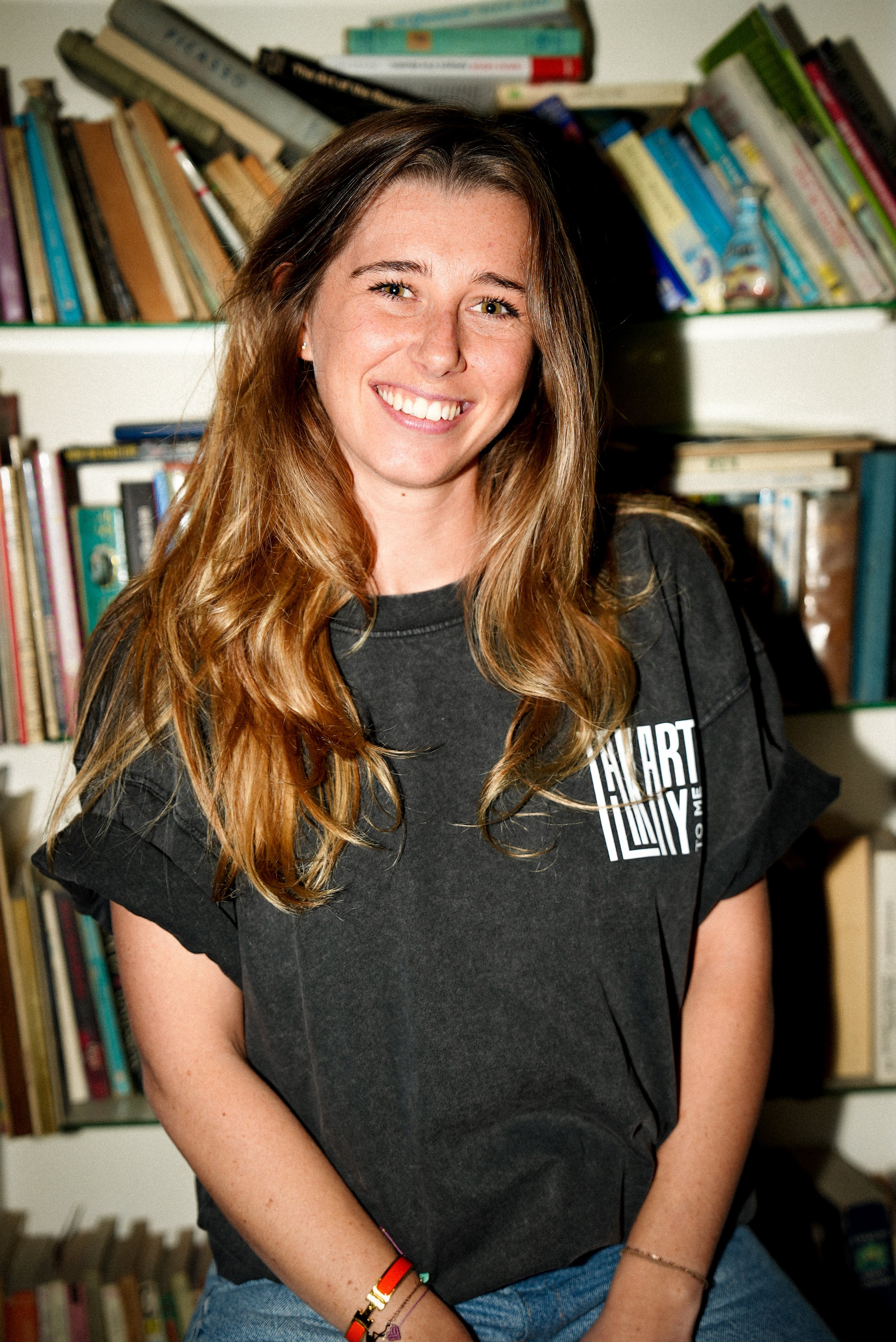 Jeune femme souriante avec des cheveux longs et châtains, portant un t-shirt noir, assise devant une étagère pleine de livres.