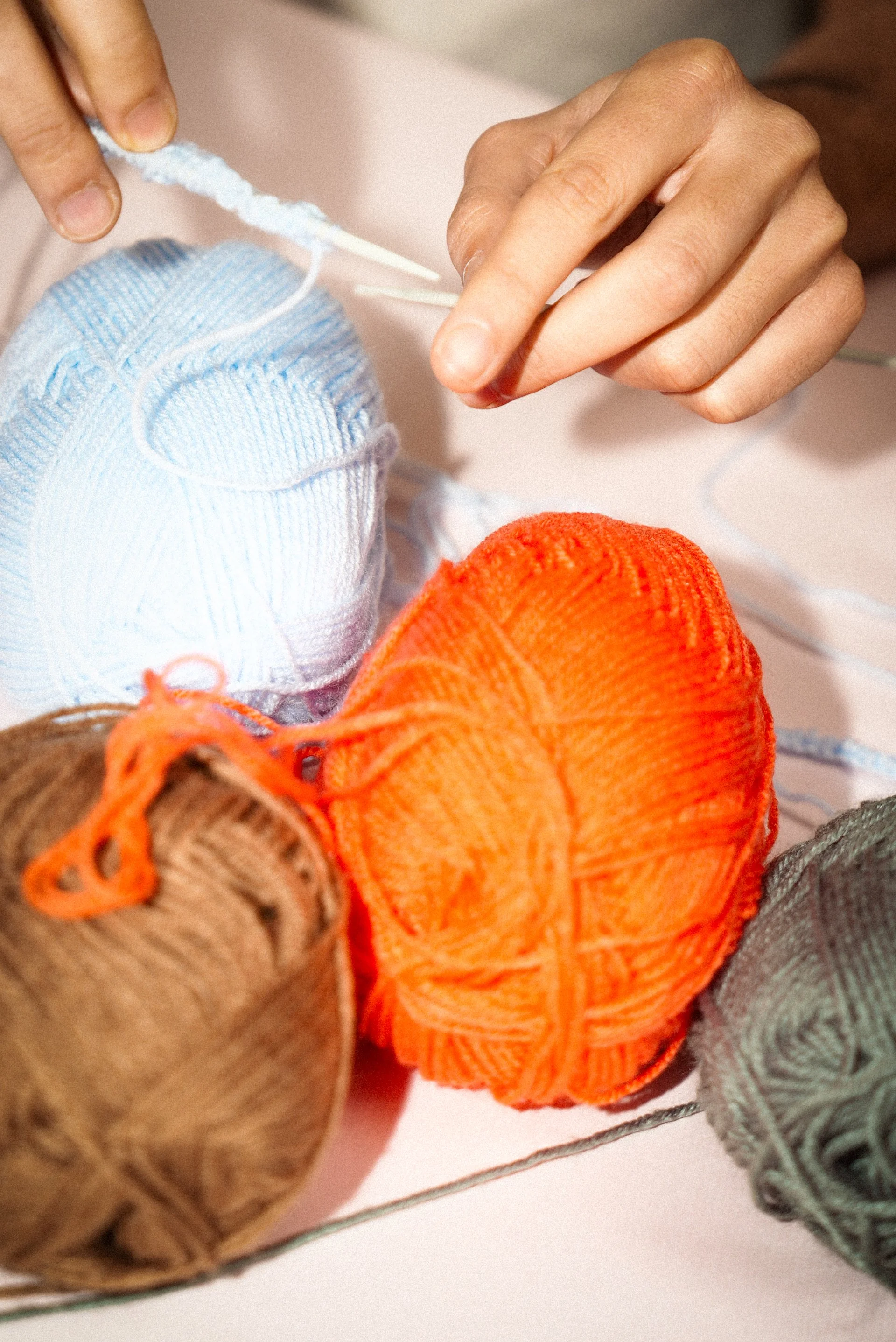 Main qui manipule une pelote de fil en coton orange, entourée de pelotes de fil de différentes couleurs (bleu, brun, gris), sur une surface claire.