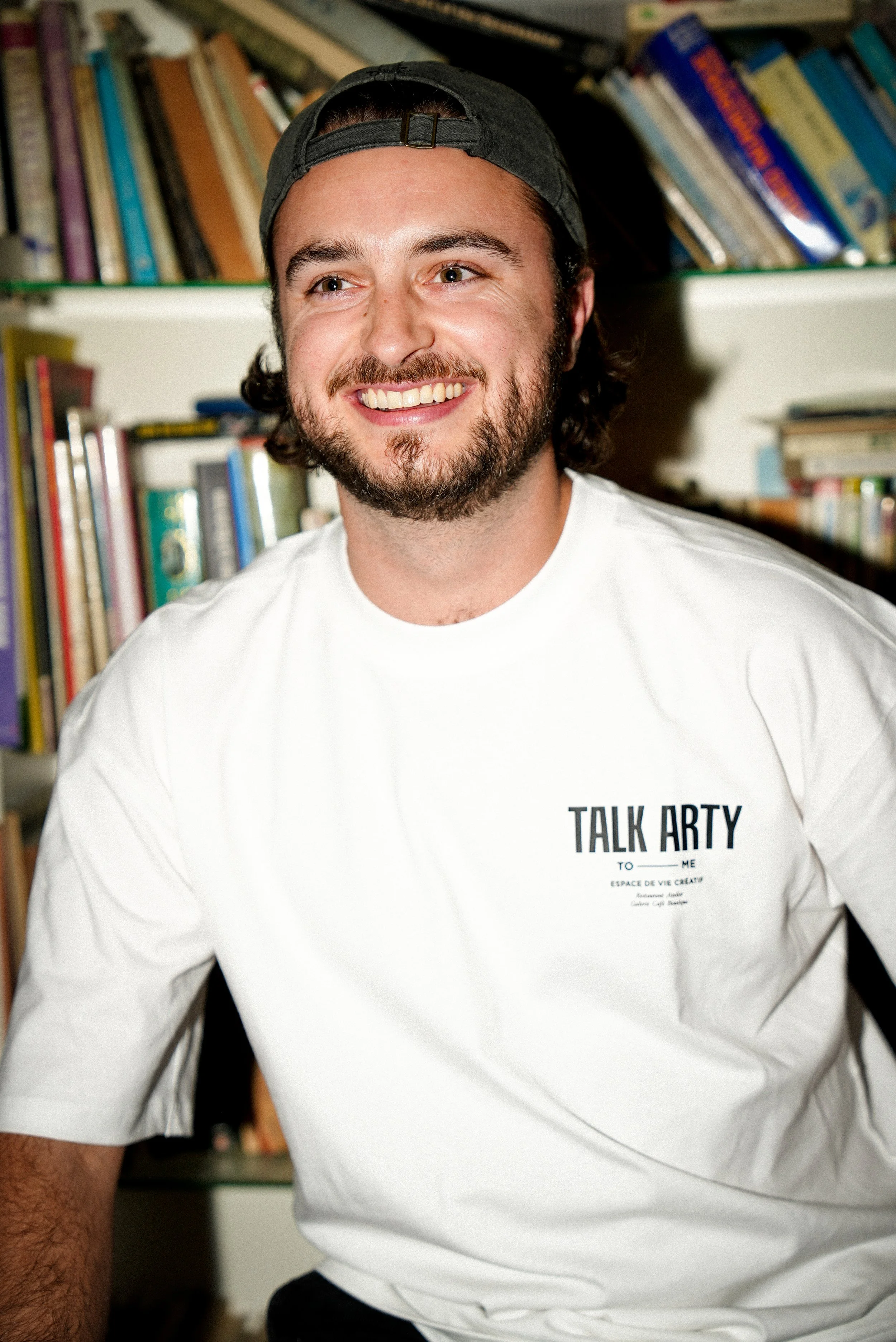 Un homme souriant portant une casquette noire, chemise blanche avec inscription 'TALK ARTY', assis devant une bibliothèque