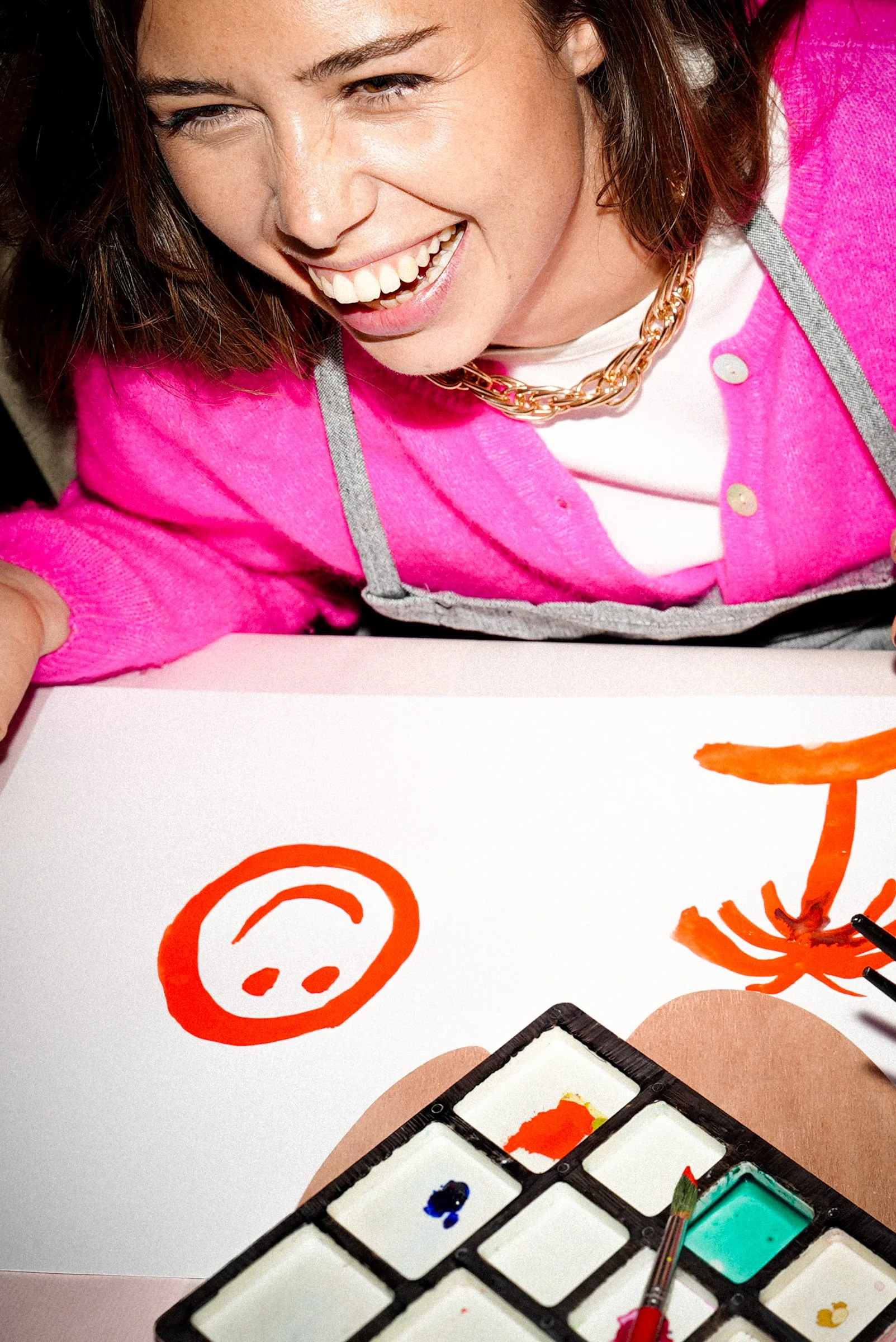 Jeune femme souriante portant un pull rose, une chaîne en or, et un tablier gris, dessinant un visage souriant en orange avec de la peinture acrylique sur une feuille. Il y a un pinceau et une palette de peinture à proximité.