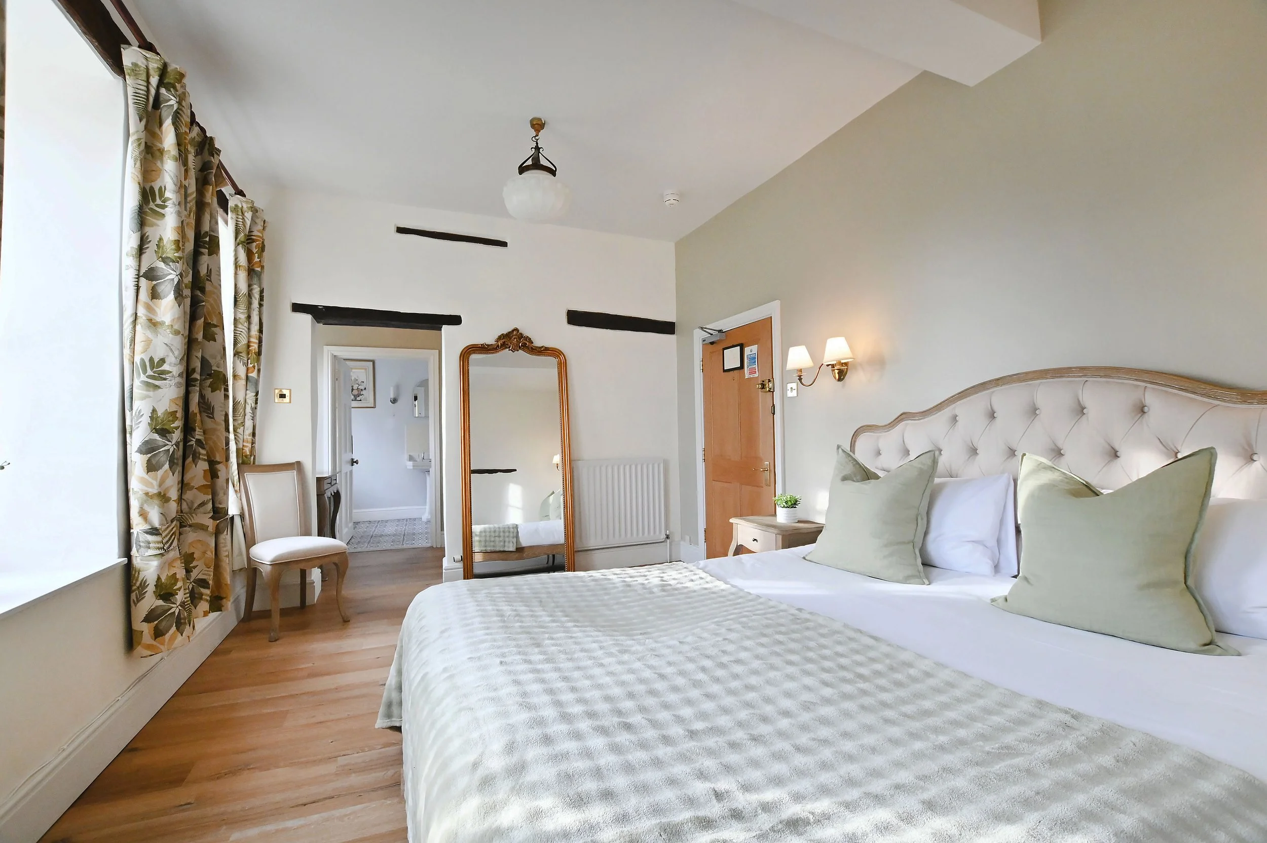 A cozy hotel bedroom with a large bed, light-colored headboard, and four pillows. A wooden nightstand with a small potted plant and wall-mounted lights is next to the bed. A walker is reflected in a tall, gold-framed mirror. Light streaming through floral-patterned curtains on a large window. A door and a chair near the window.