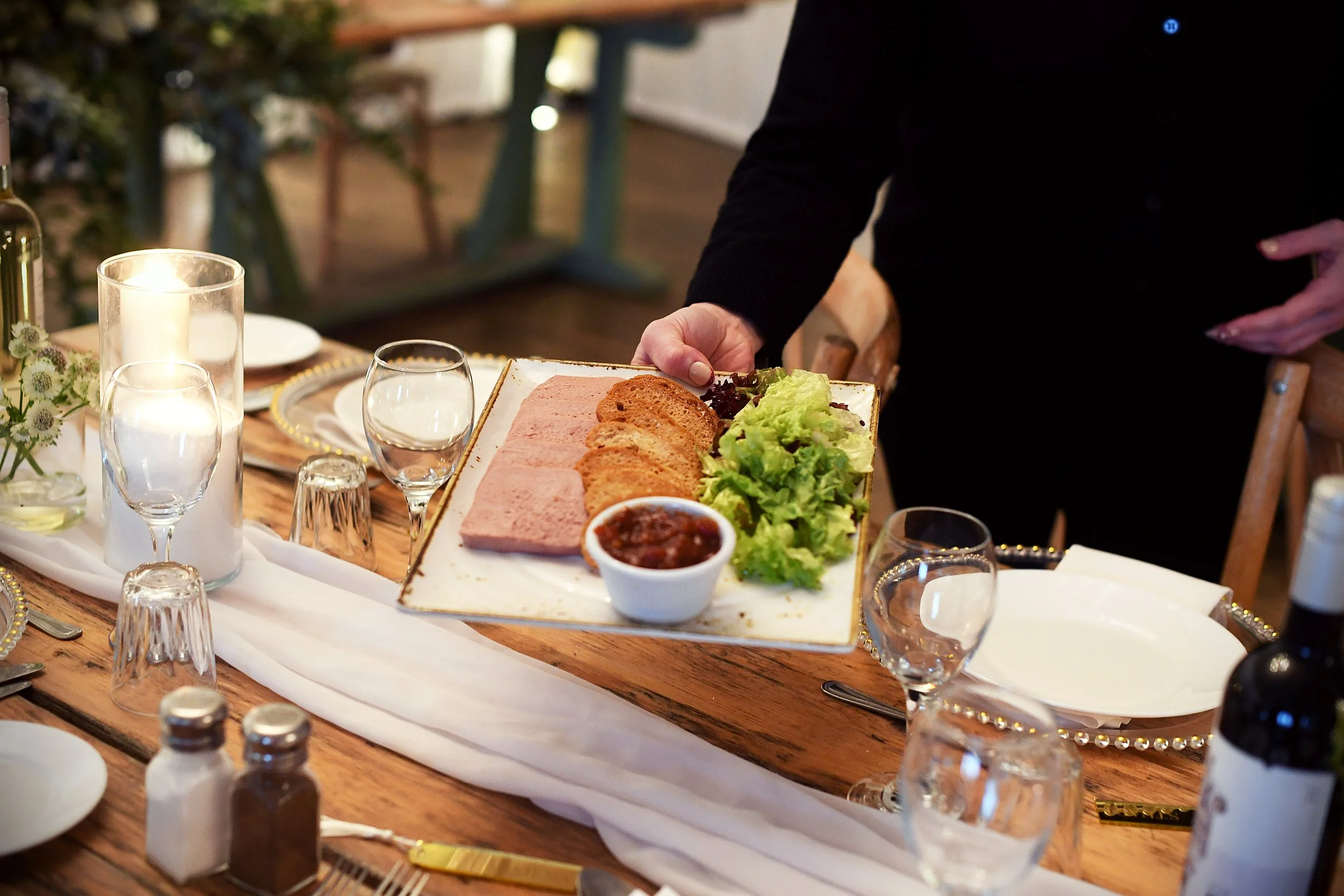 Person serving a rectangular appetizer platter with sliced bread, leafy greens, and a bowl of chili at a dinner table.