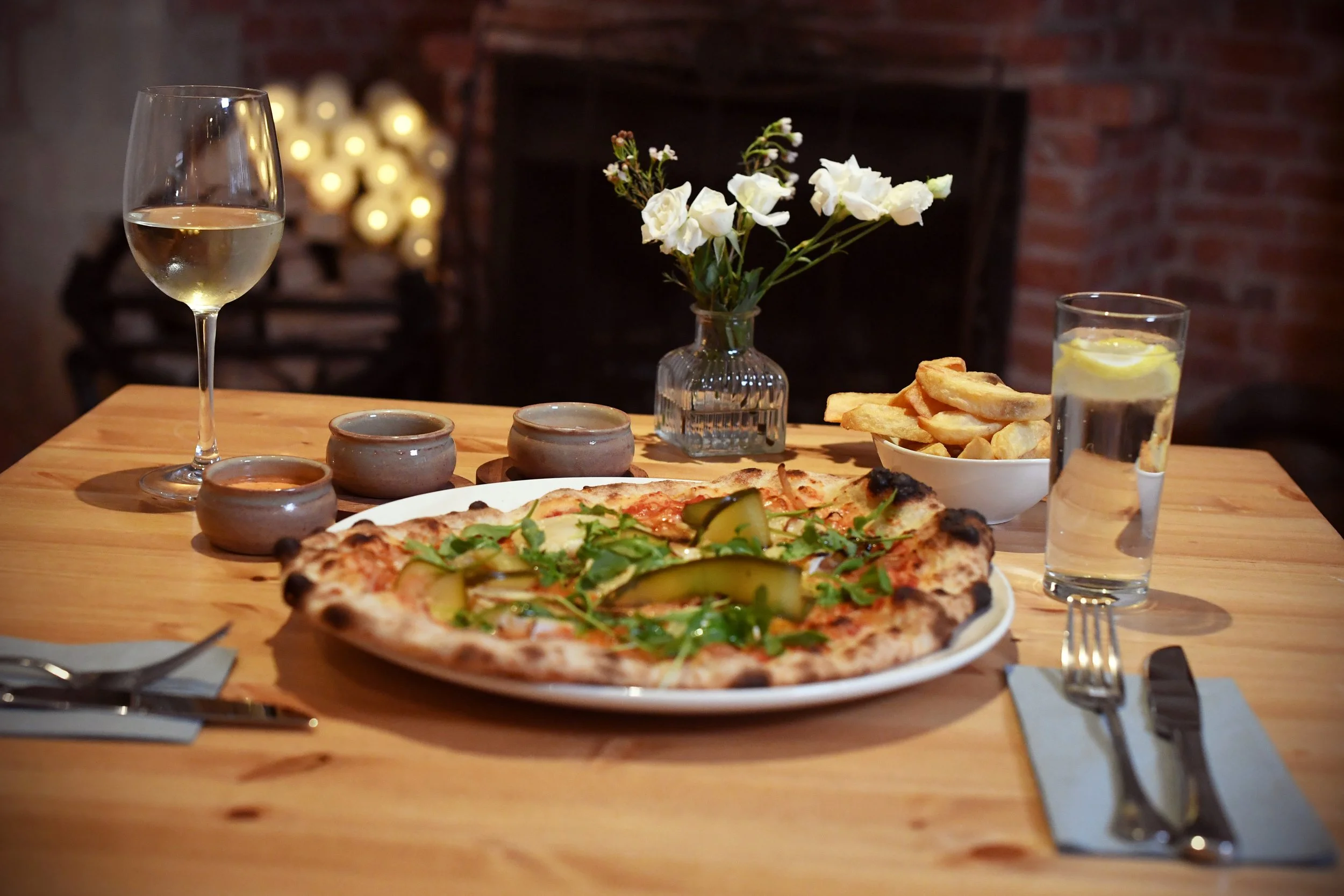 A wooden table set for a meal with a pizza topped with zucchini and greens, a glass of white wine, a glass of water with lemon, a bowl of potato fries, a small vase with white flowers, three small bowls, and utensils on napkins.