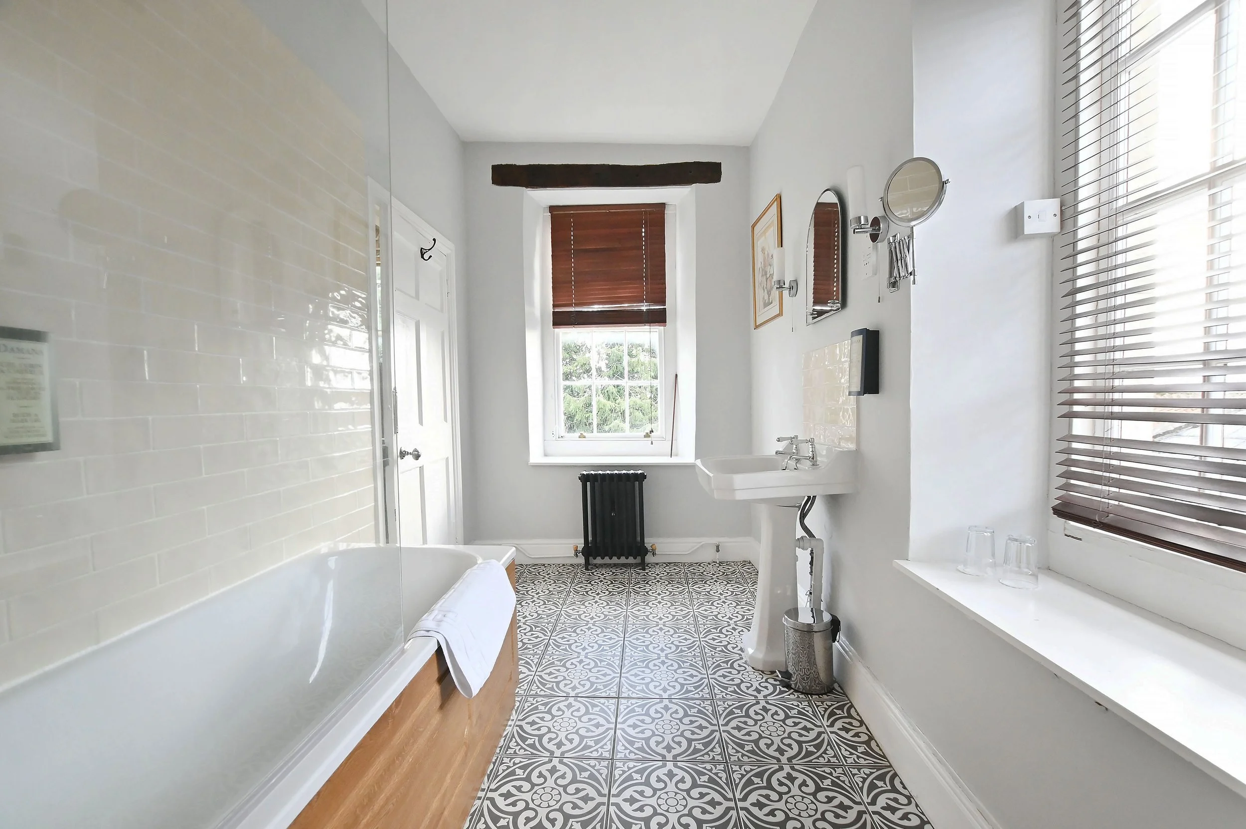 Bright bathroom with patterned floor tiles, white bathtub, pedestal sink, window with brown blinds, wall mirrors, and various small items.
