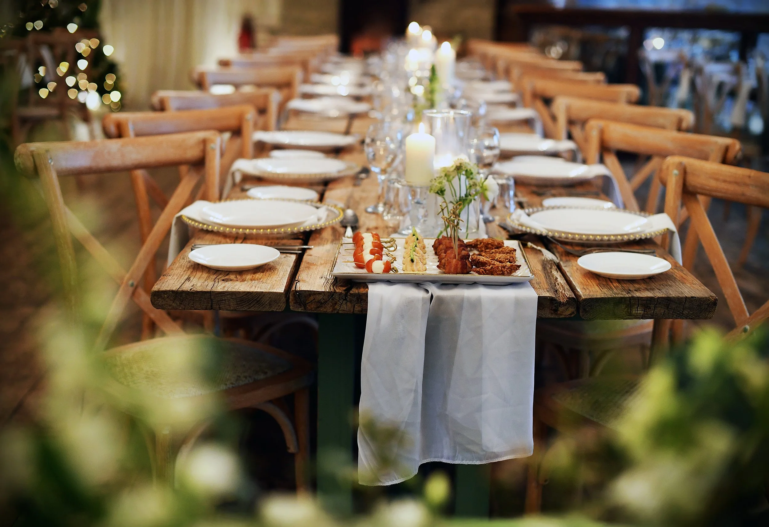 A rustic outdoor dining table set for a special occasion, with a white tablecloth draped over the center, surrounded by wooden chairs, and decorated with candles, flowers, and a tray of appetizers.