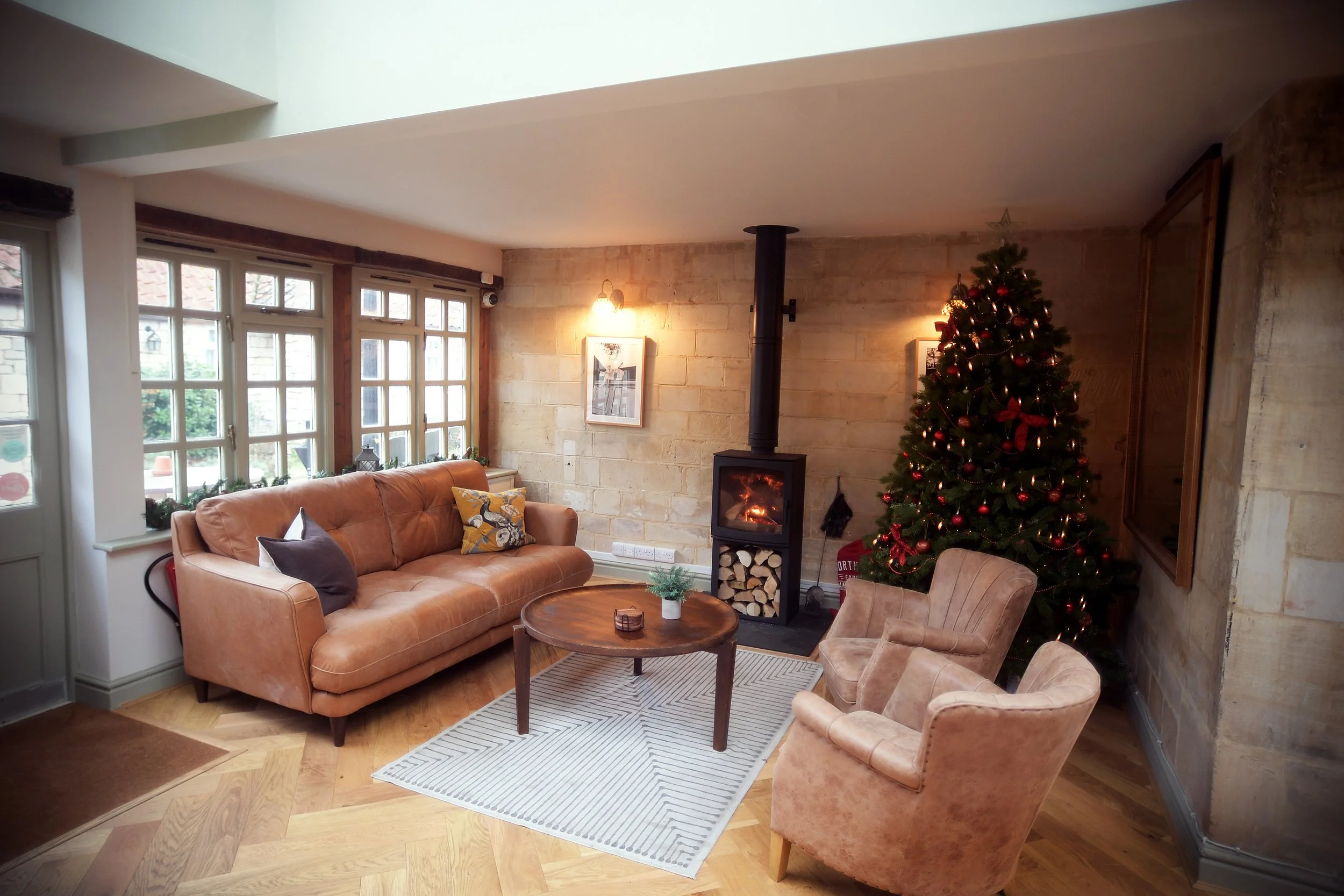 Living room decorated for Christmas with a Christmas tree, fireplace, leather sofa, two armchairs, wooden coffee table, and windows in the background.