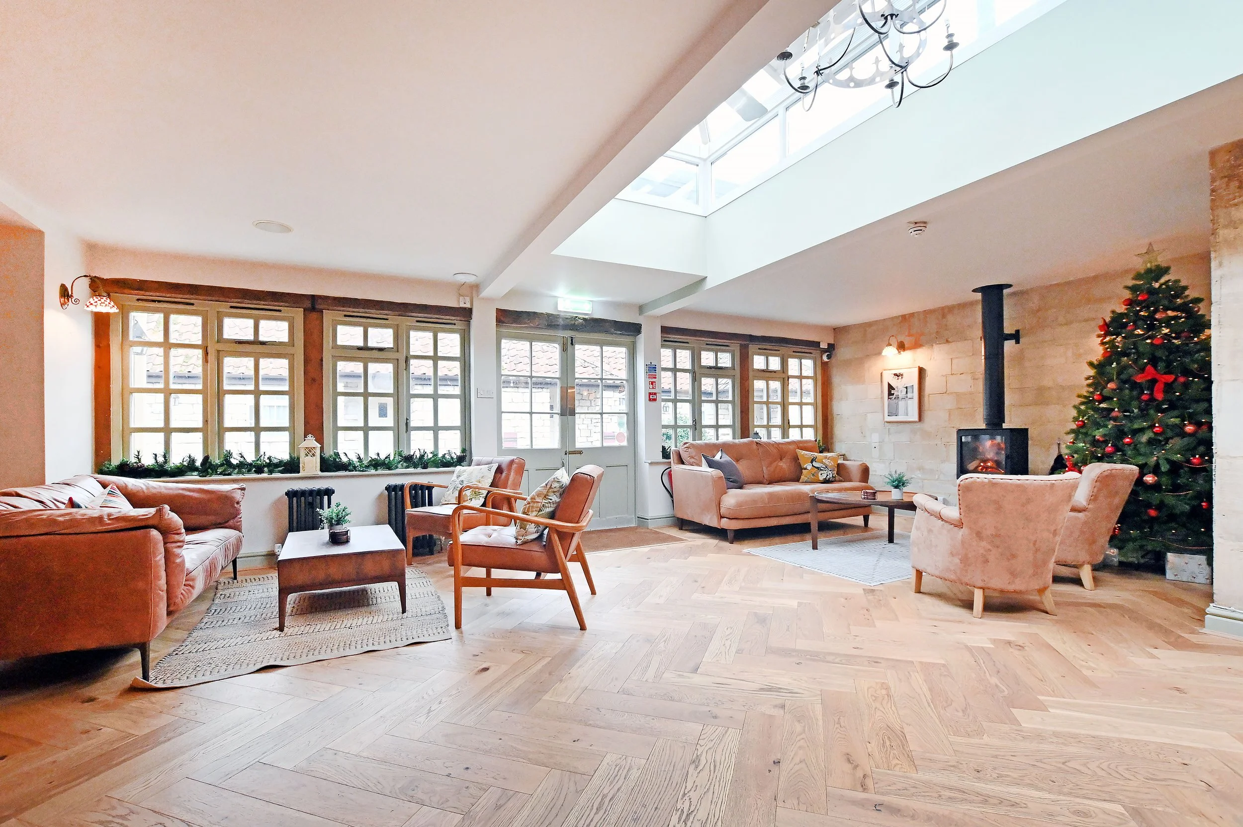 A cozy living room decorated for Christmas with a large Christmas tree, leather sofas, armchairs, a wood stove, and large windows letting in natural light.